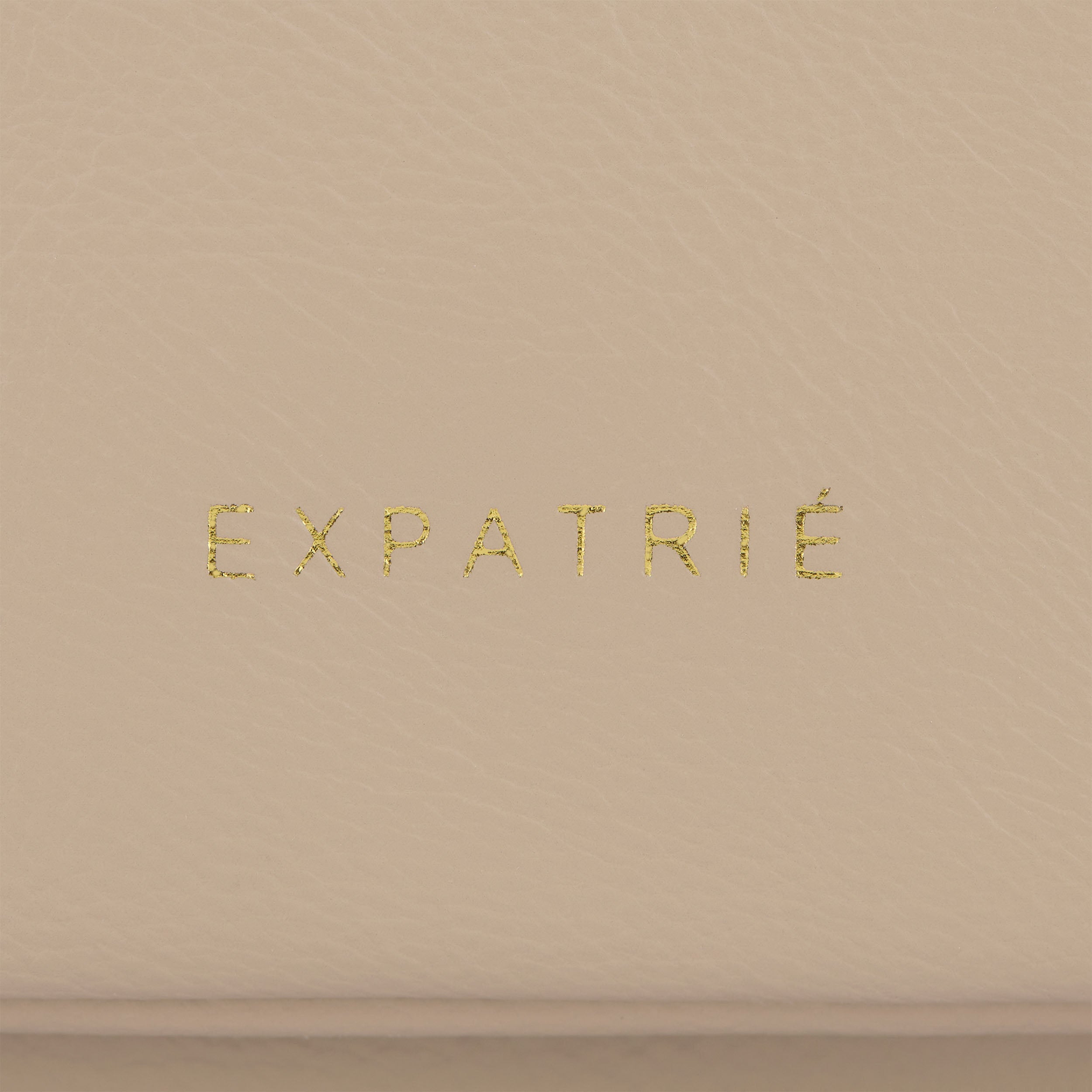 Beige Kunstleder-Oberfläche mit goldenem Schriftzug 'EXPATRIÉ' auf Clutch Juliette