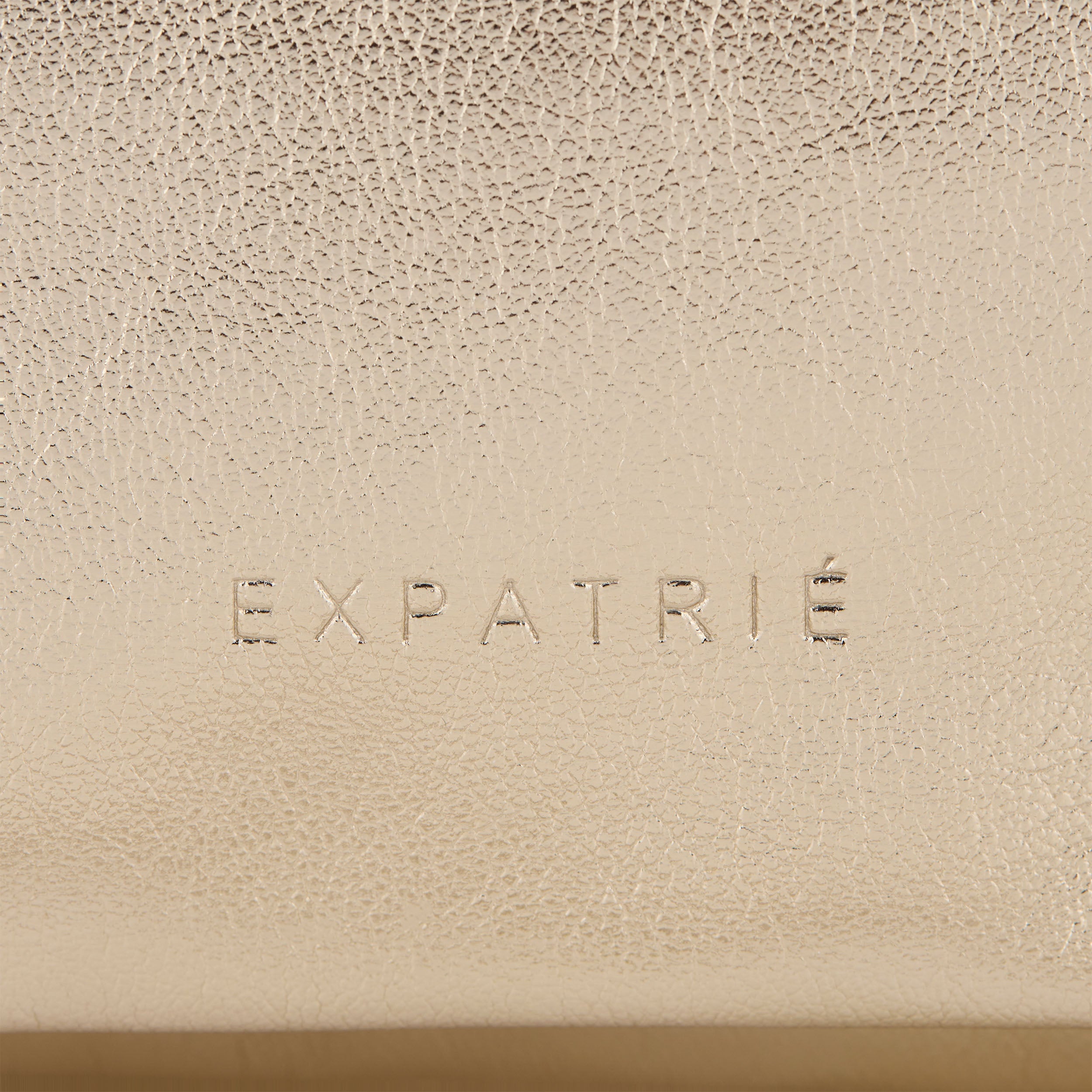 Nahaufnahme der geprägten Marke 'EXPATRIÉ' auf goldfarbenem Kunstleder der Clutch Juliette