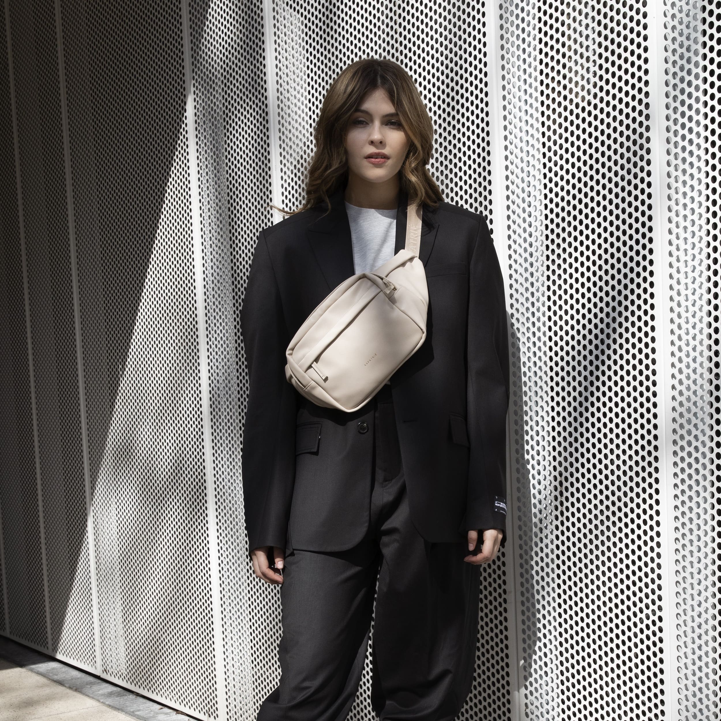 Modell trägt beige Bauchtasche Justine PU über schwarzen Blazer und Hose vor perforiertem Metallhintergrund