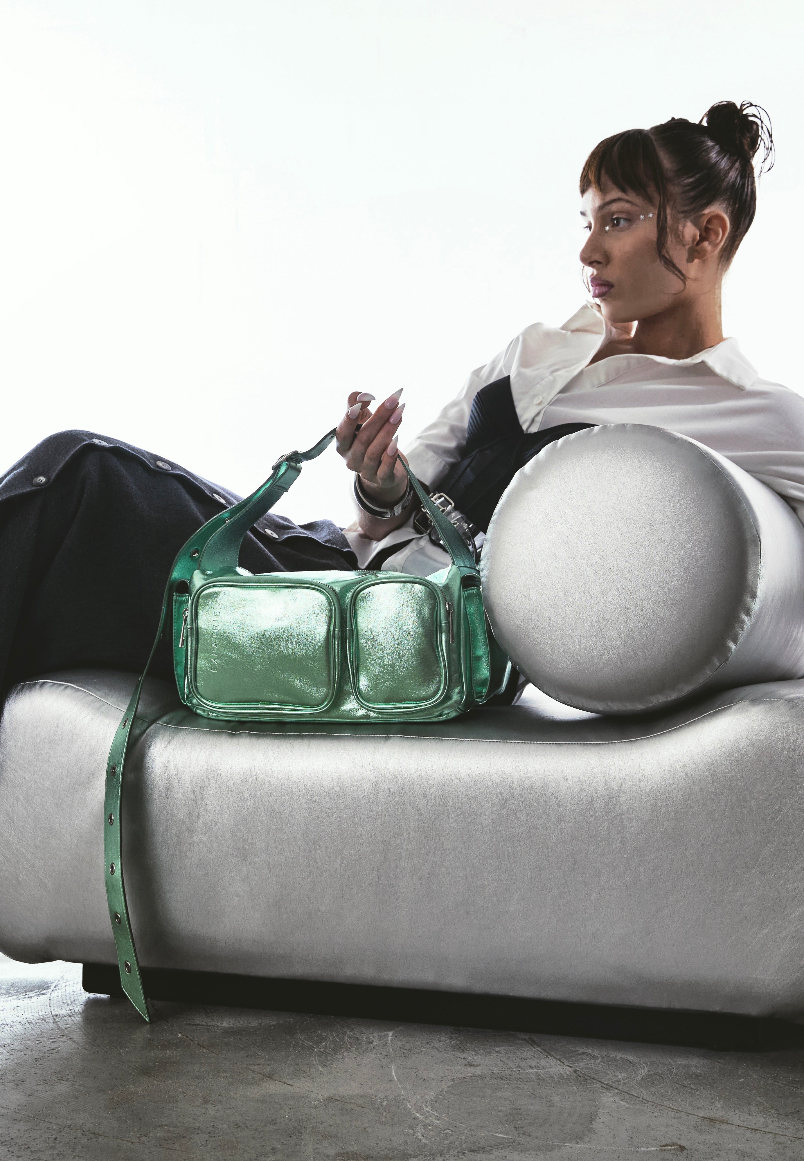 Grüne STELLA Schultertasche mit zwei Frontfächern aus Leder auf silberner Couch neben Frau in weißem Hemd und schwarzem Rock