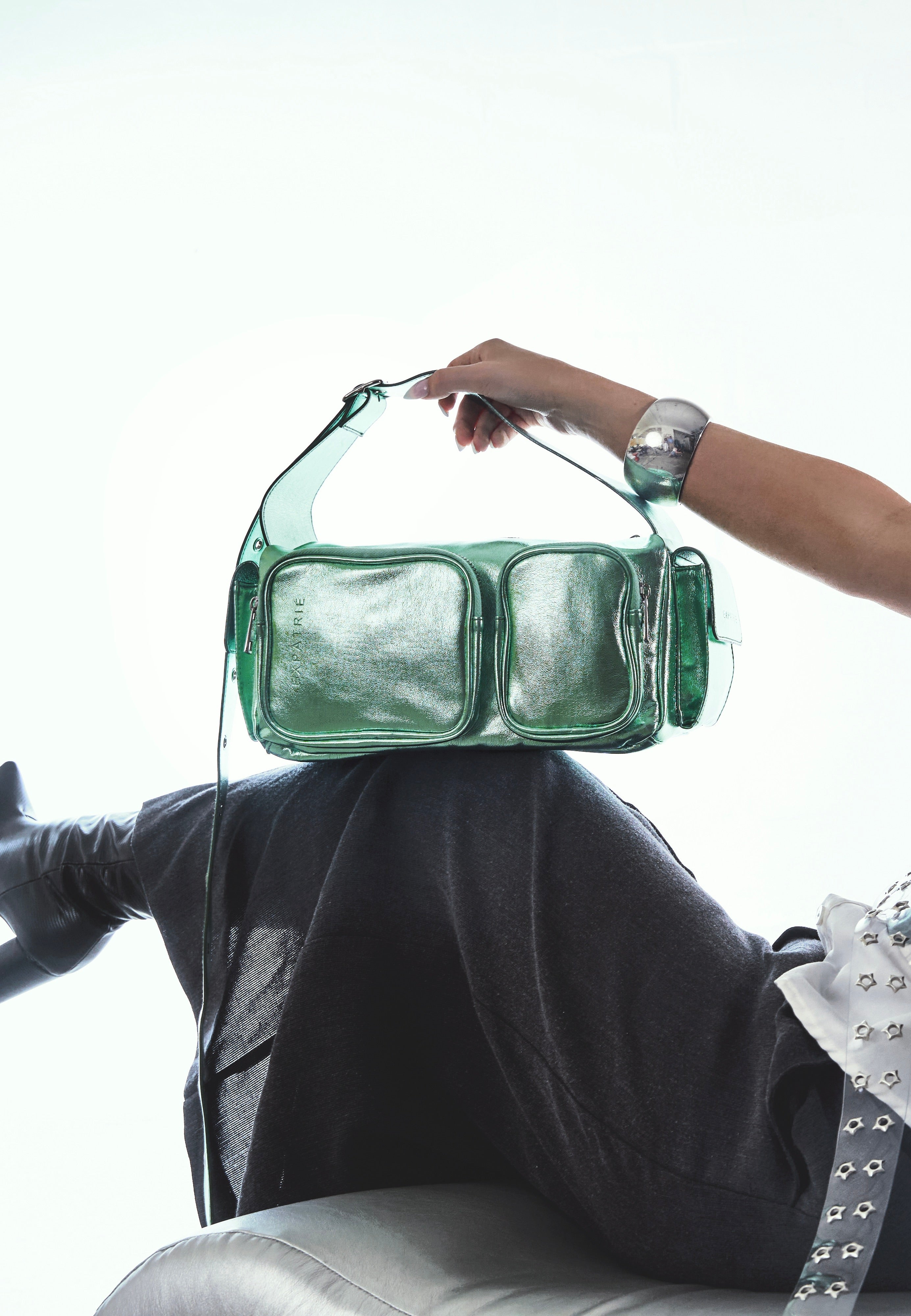 Grüne STELLA Schultertasche aus glänzendem Material mit zwei Außentaschen, gehalten von einer Person mit silbernem Armreif