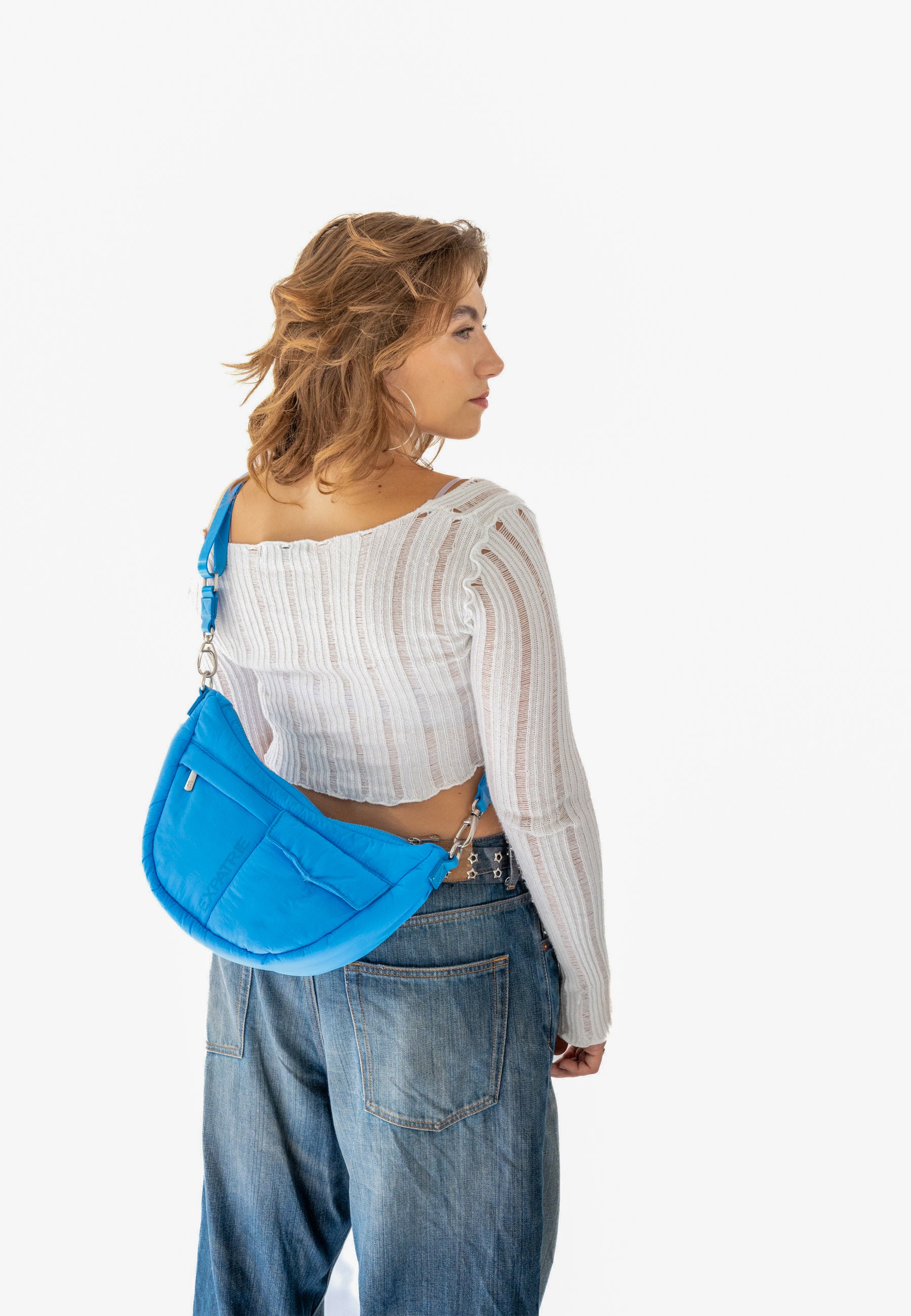 Frau mit lockigem Haar trägt blaue VIVY Half Moon Bag lässig über der Schulter, dazu weißes, langärmliges Top und blaue Jeans