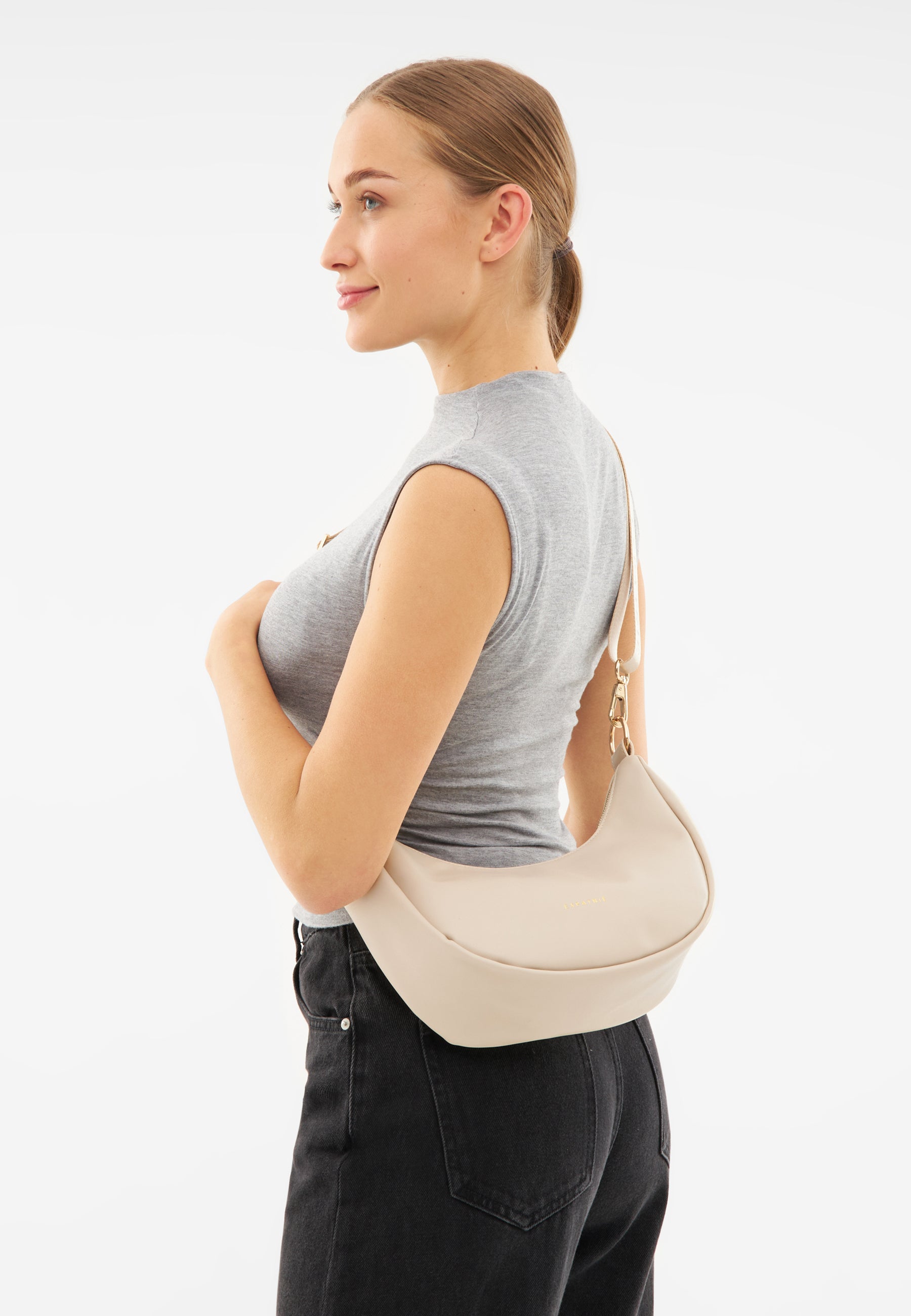Frau mit beiger LEA SMALL Half Moon Bag Over-Shoulder, graues ärmelloses Oberteil, schwarze Jeans