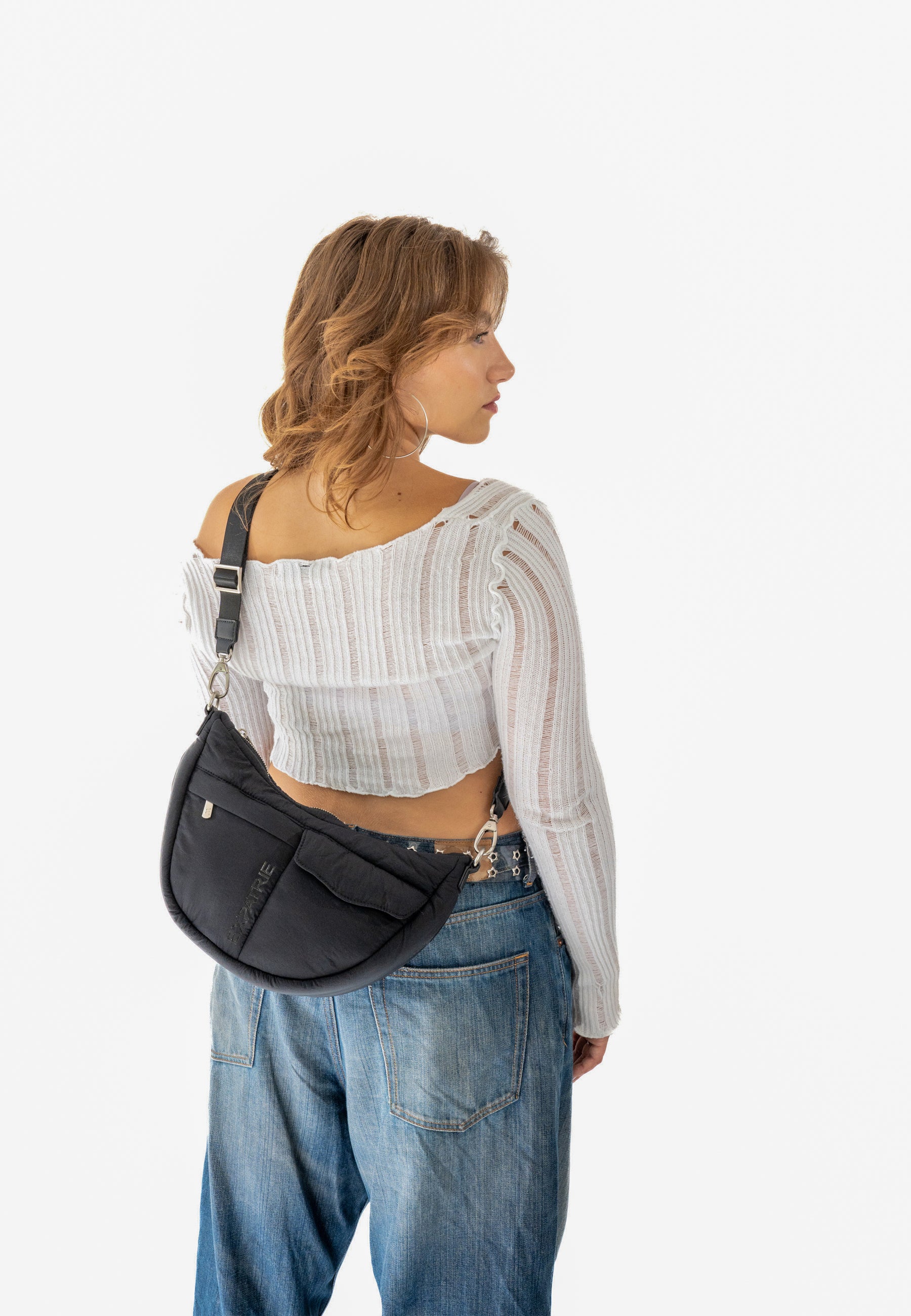 Frau trägt VIVY Half Moon Bag in Schwarz quer über den Rücken mit weißem, langärmligem, geripptem Crop-Top und blauen Jeans