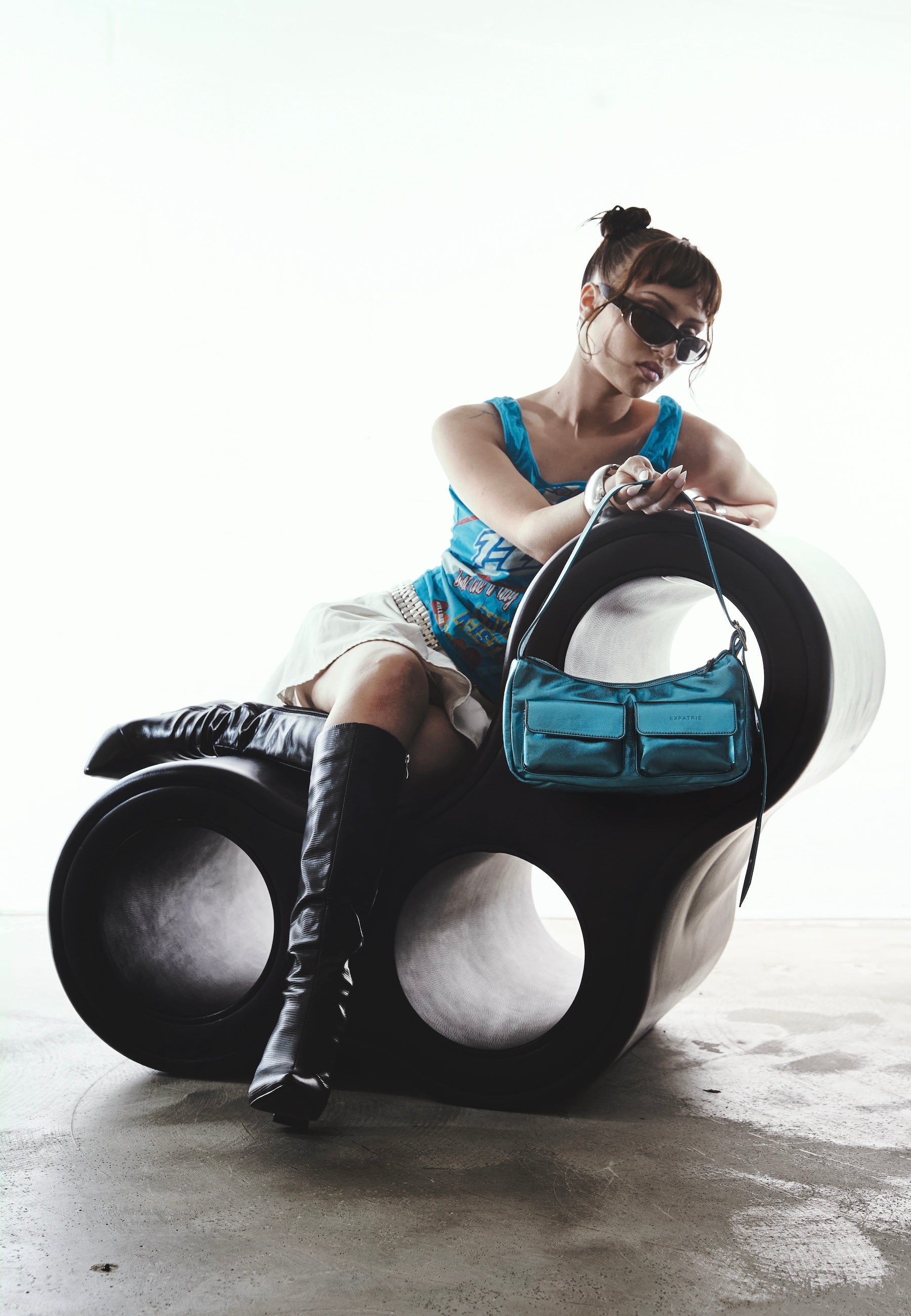 Frau mit schwarzen Stiefeln und blauem Top sitzt auf modernem schwarzen Sitzobjekt, hält blaue metallic Handtasche ELECTRA