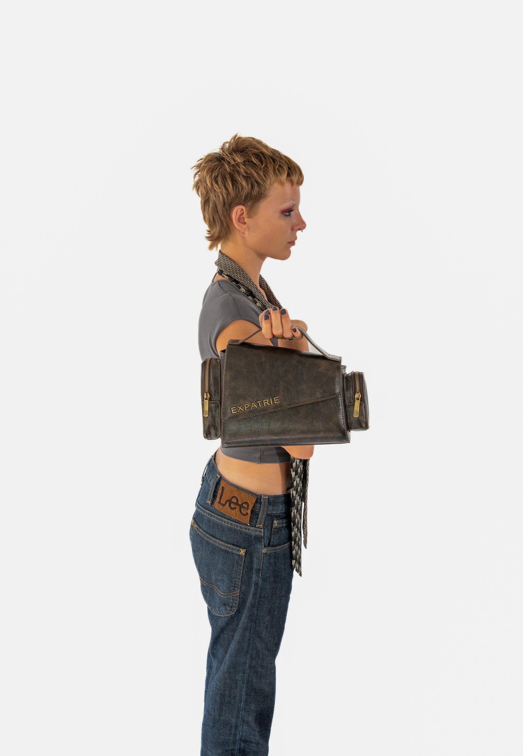 Frau mit kurzem Haar trägt graues Oberteil und Jeans, hält HUNTER Handtasche in Braun vintage mit EXPRATIE Schriftzug seitlich