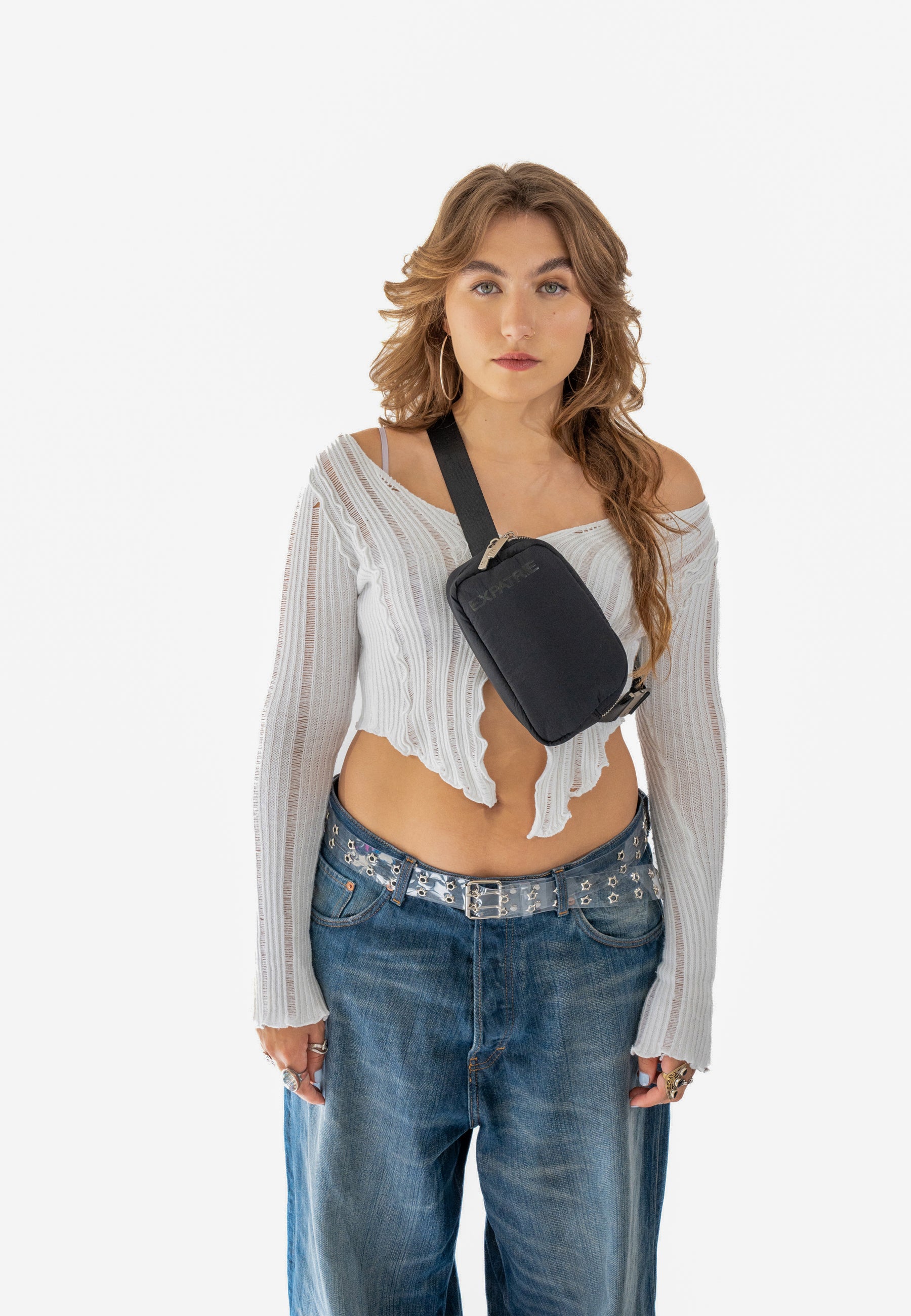 Frau mit weißem, langärmligem, gerippten Crop-Top und ausgestellter Jeans trägt schwarze MILLY Bauchtasche quer über die Brust
