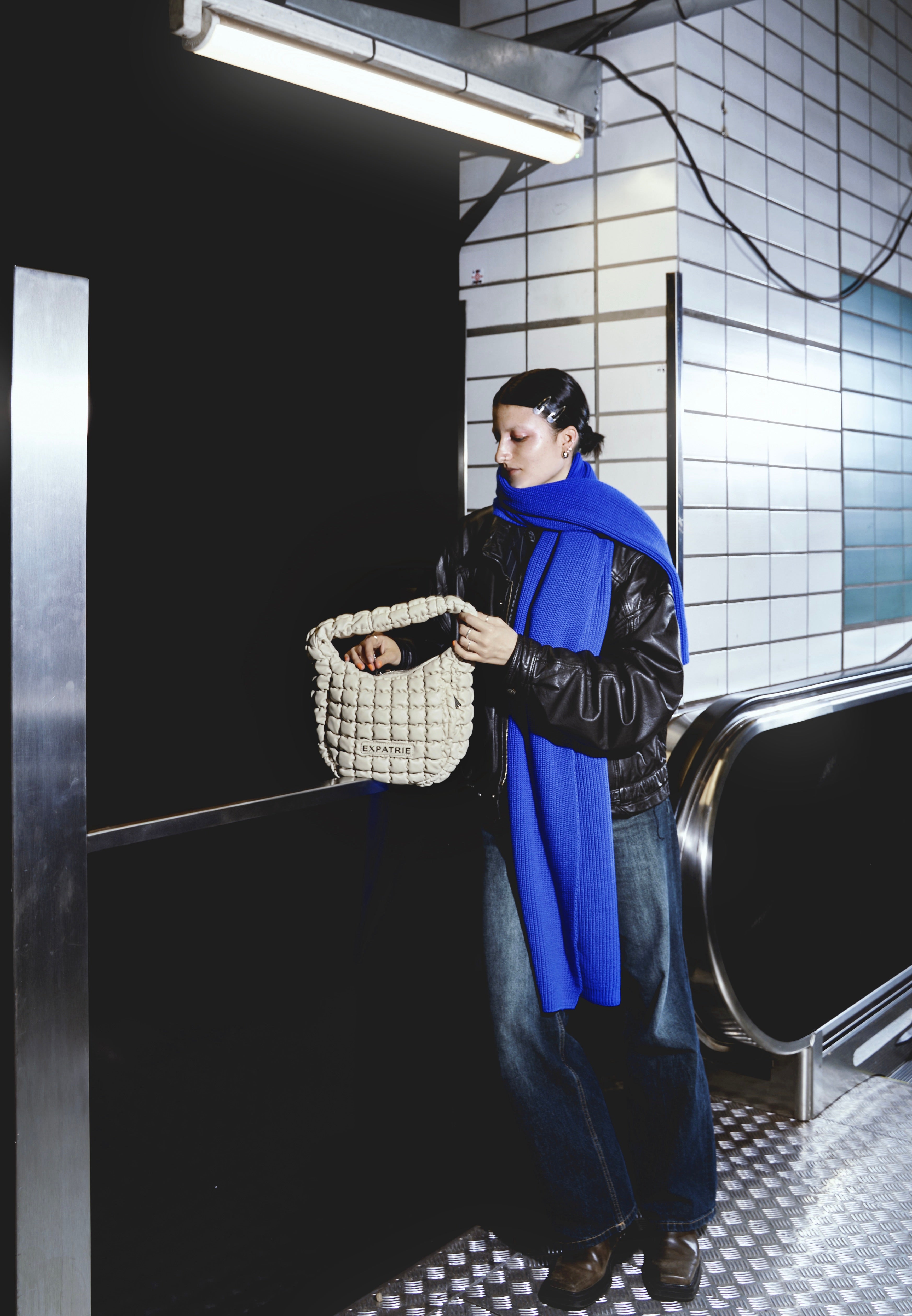 Frau in Lederjacke und blauem Schal hält beigetöne PIPPA LARGE Handtasche in U-Bahn-Station