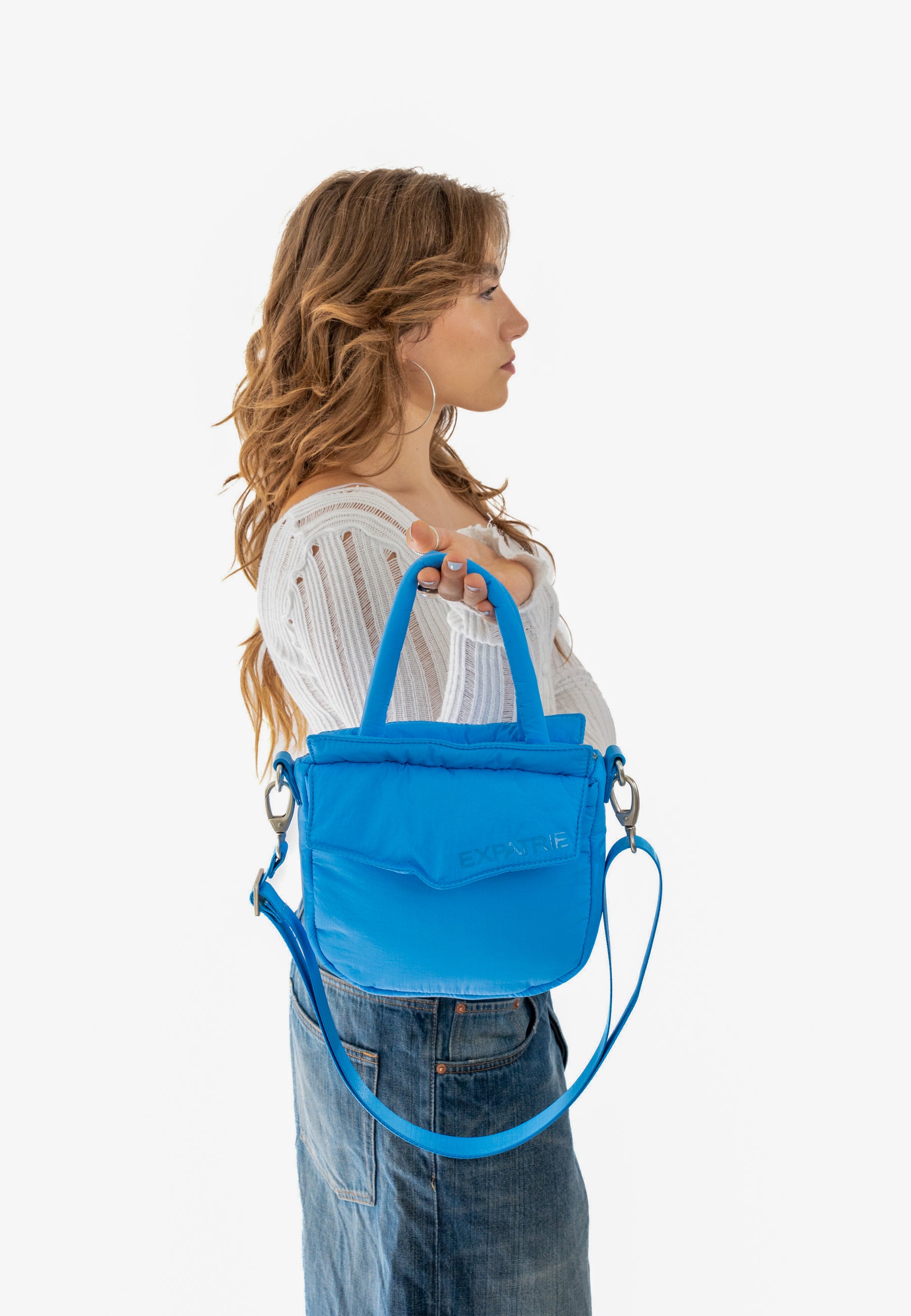 Frau zeigt blaue POPPY SMALL Handtasche mit Griffen und Schulterriemen