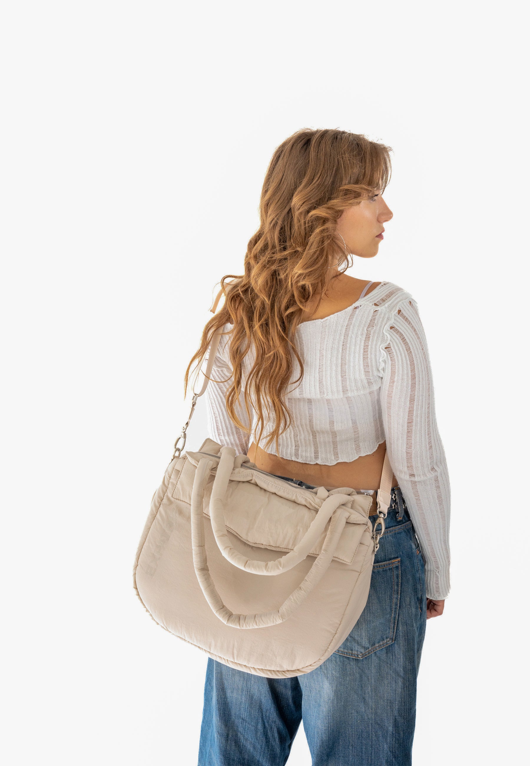 Frau trägt beige gepolsterte POPPY Shopper Tasche über Schulter zu weißem, durchsichtigem Oberteil und blauen Jeans