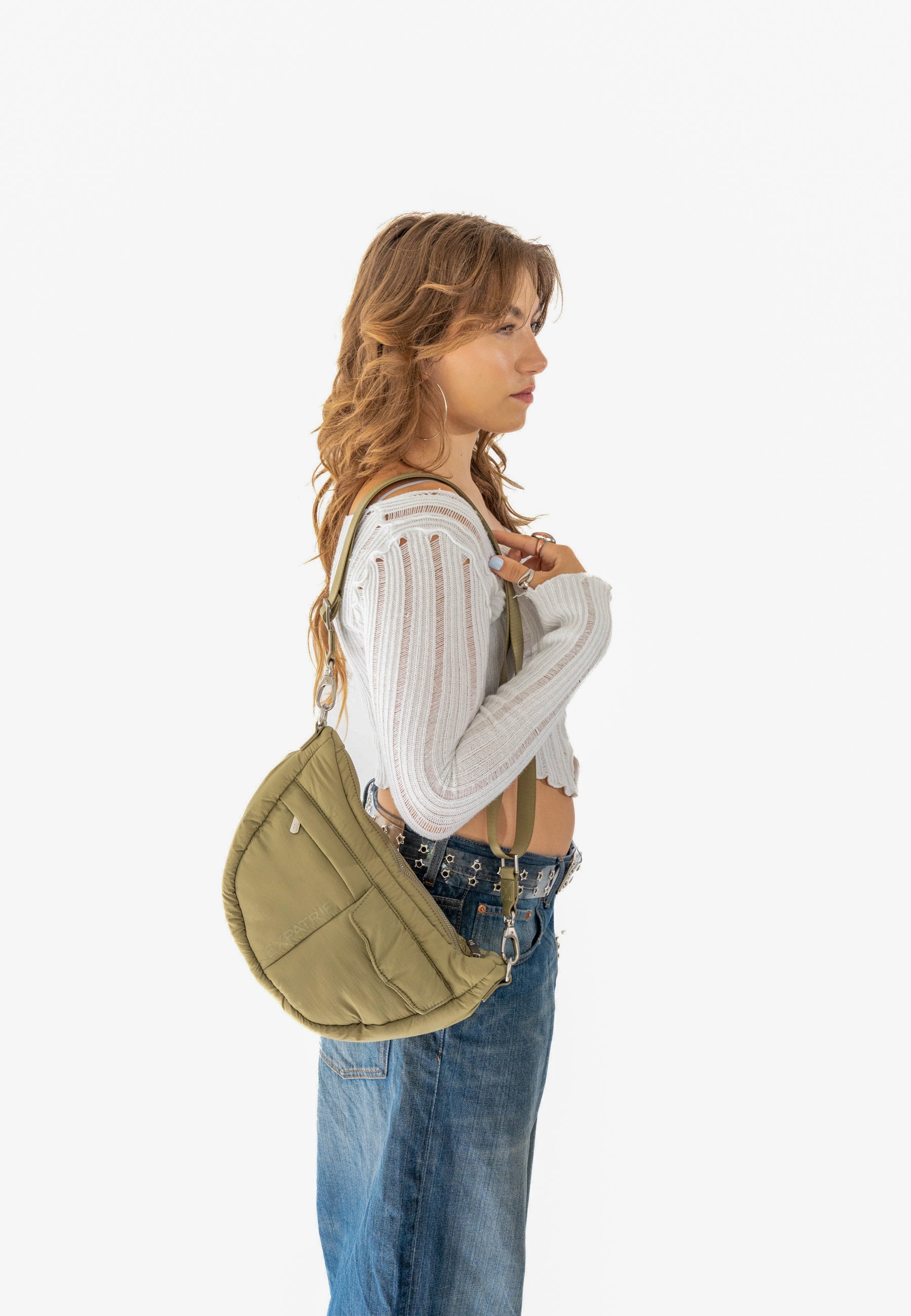 Frau von der Seite mit langen, gewellten Haaren, trägt eine olivgrüne VIVY Half Moon Bag und lässiges Outfit mit weißem, langärmeligen Oberteil und Jeansrock