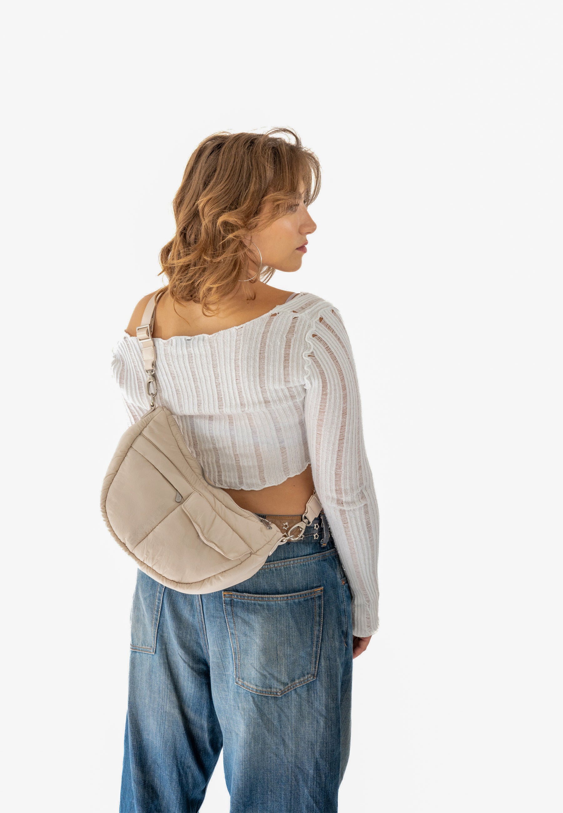 Frau trägt beige VIVY Half Moon Bag über dem Rücken mit weißem, langärmligem, transparentem Oberteil und blauen Jeans