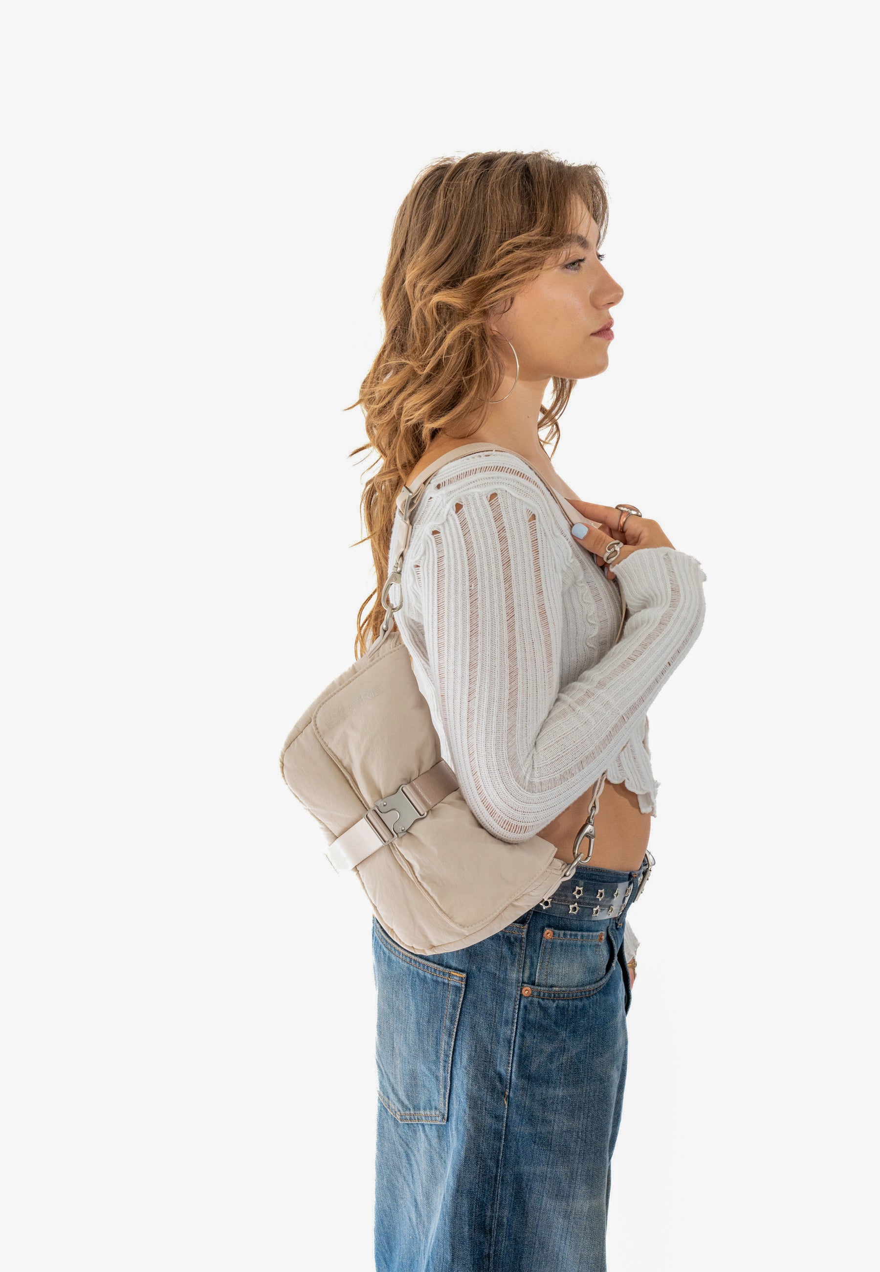Seitenansicht einer Frau mit lockigen Haaren, die eine beige BOBBY Handtasche aus gepolstertem Material über der Schulter trägt, dazu weißes, durchsichtiges Rippenstrick-Top und blaue Jeans mit Gürtel