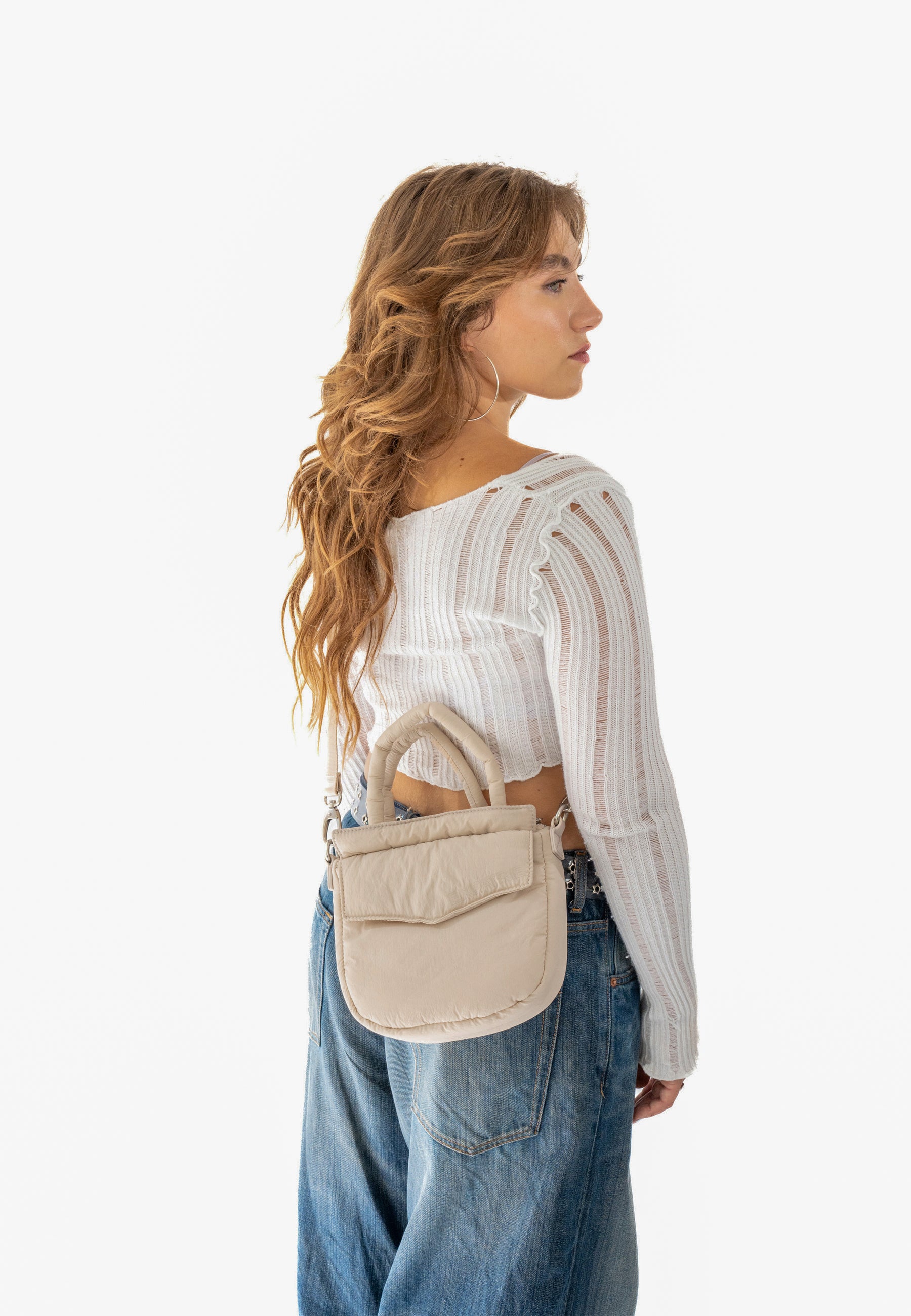 Frau mit langen braunen Haaren, die beige gepolsterte Tasche 'POPPY SMALL' trägt, kombiniert mit weißem, langärmligem, durchscheinendem Oberteil und blauen Jeans