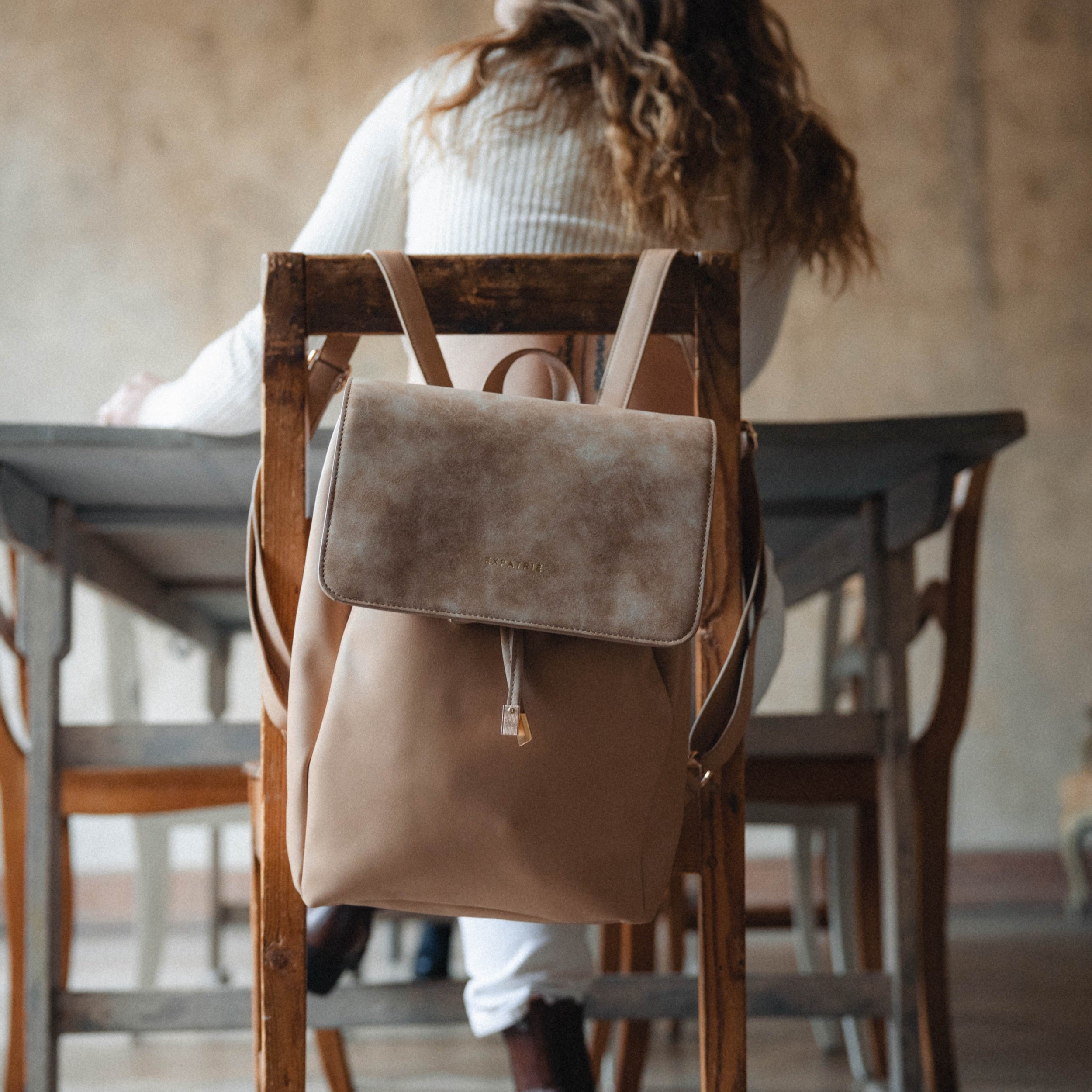 Beiger Kunstleder Rucksack Estelle hängt an einem Holzstuhl, Frau sitzt am Tisch im Hintergrund