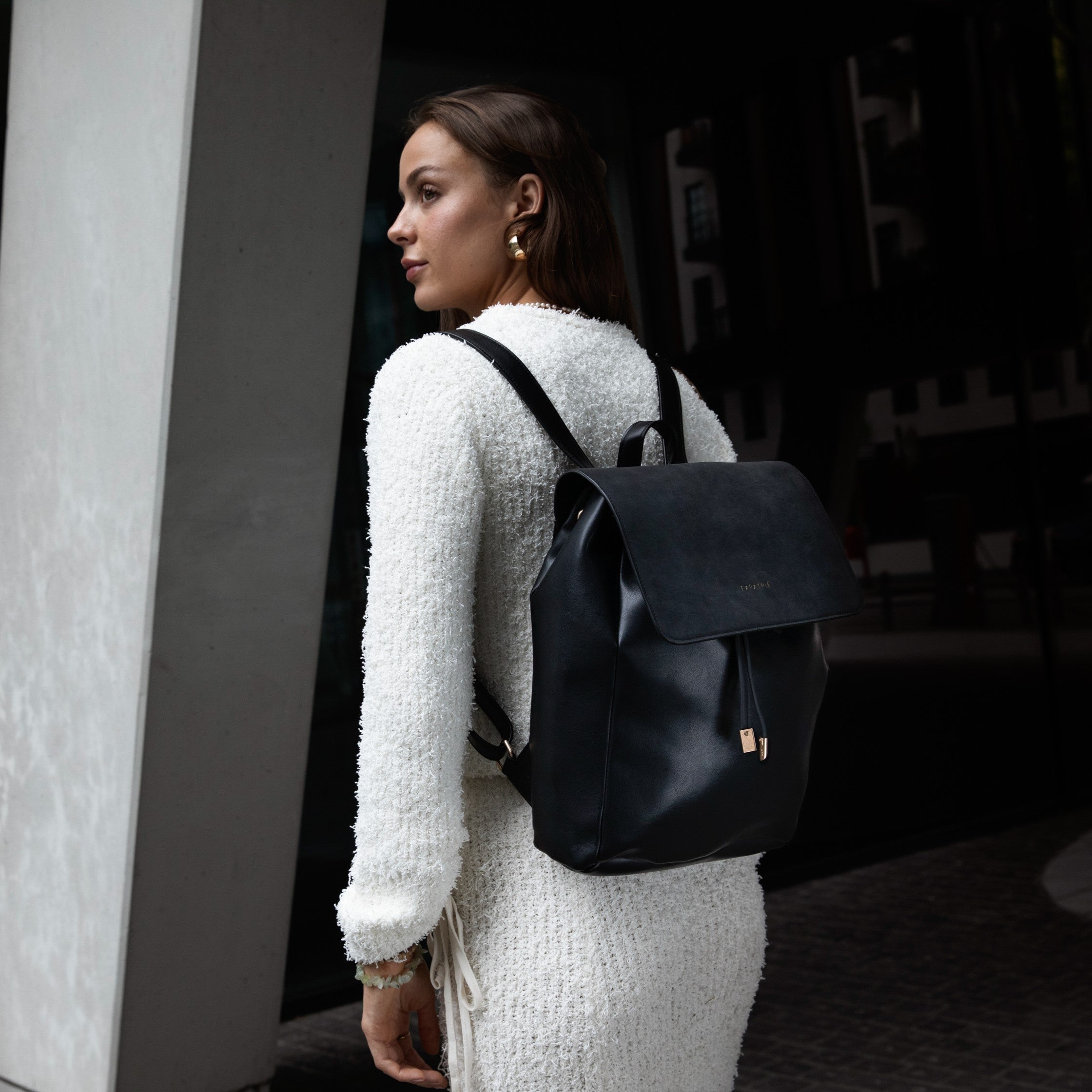 Frau trägt schwarzen Kunstleder-Rucksack Estelle mit Klappe zu cremeweißem, strukturiertem Kleid