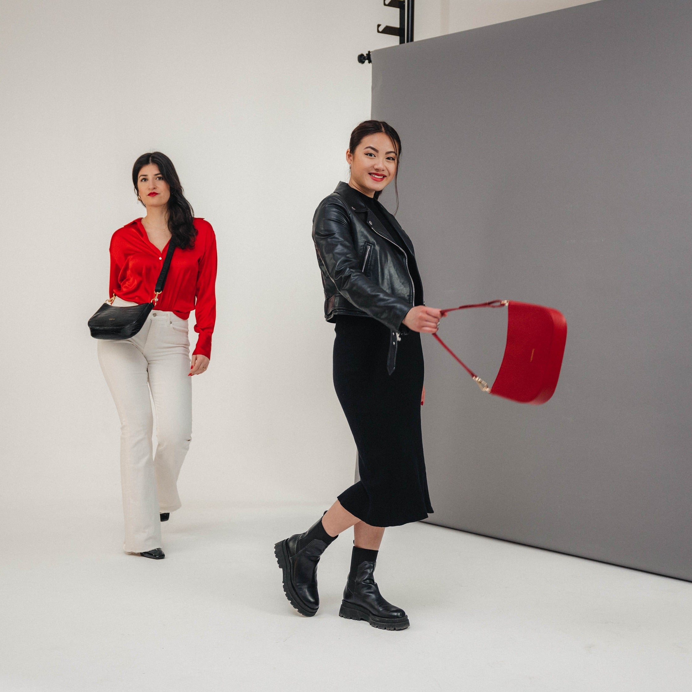 Frau mit roter Handtasche Féline, schwarze Lederjacke, Kleid und Stiefel, daneben Frau mit schwarzer Umhängetasche, roter Bluse und weißen Hosen