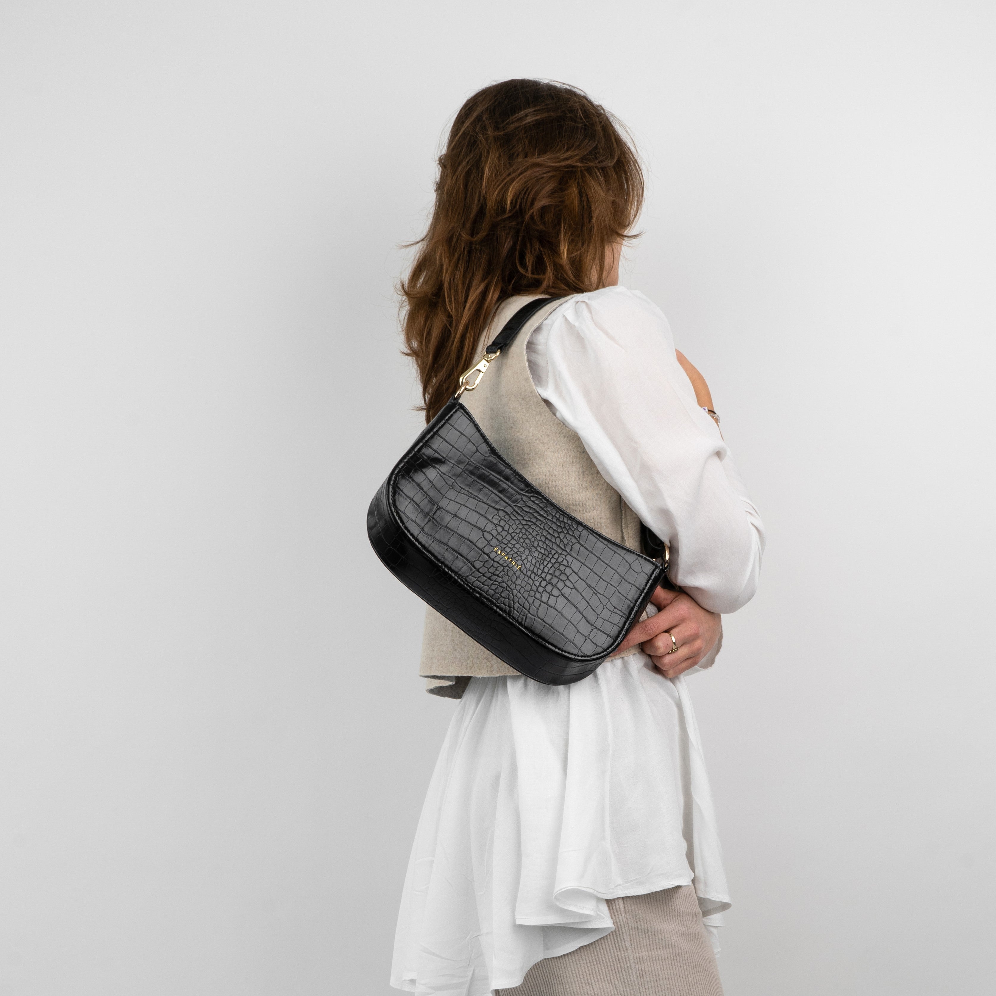 Schwarze Schultertasche 'Féline' mit Krokodilprägung an Frau in weißem Oberteil und beige Weste