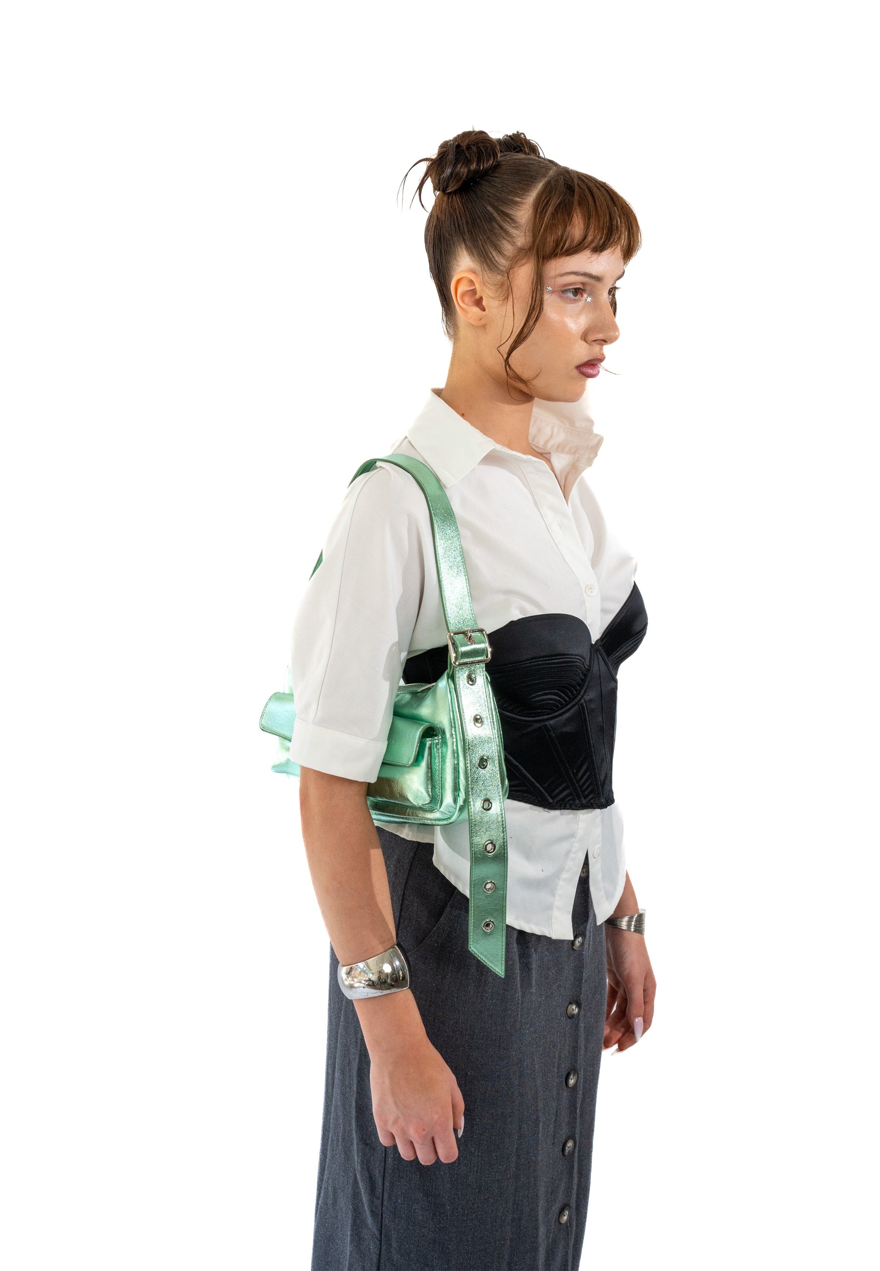 Frau mit weißer Bluse und schwarzem Korsett trägt grüne metallic ‚Electra‘ Handtasche über der Schulter