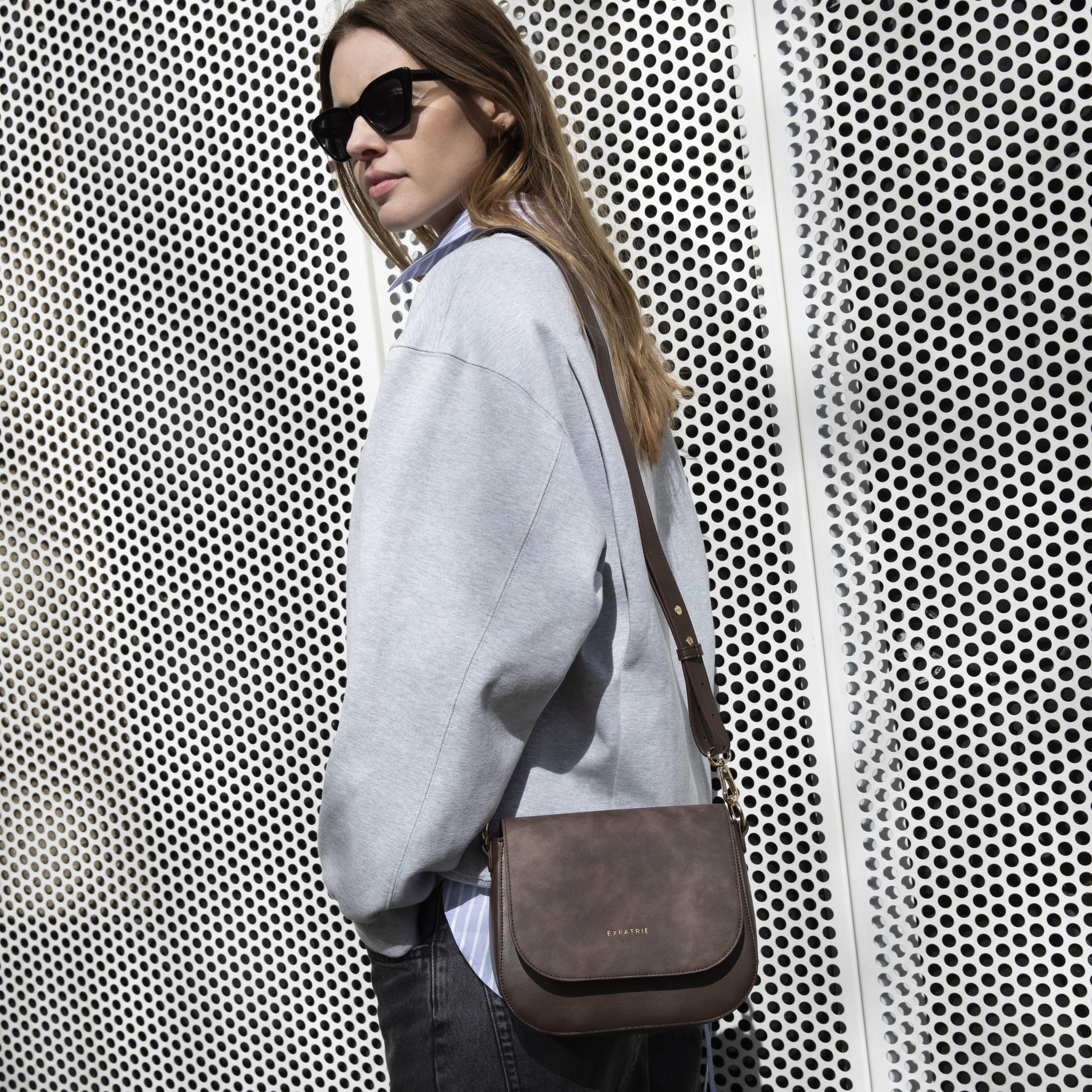 Frau mit Sonnenbrille trägt dunkelbraune Expatrié Saddle Bag Louise Medium Umhängetasche aus Leder