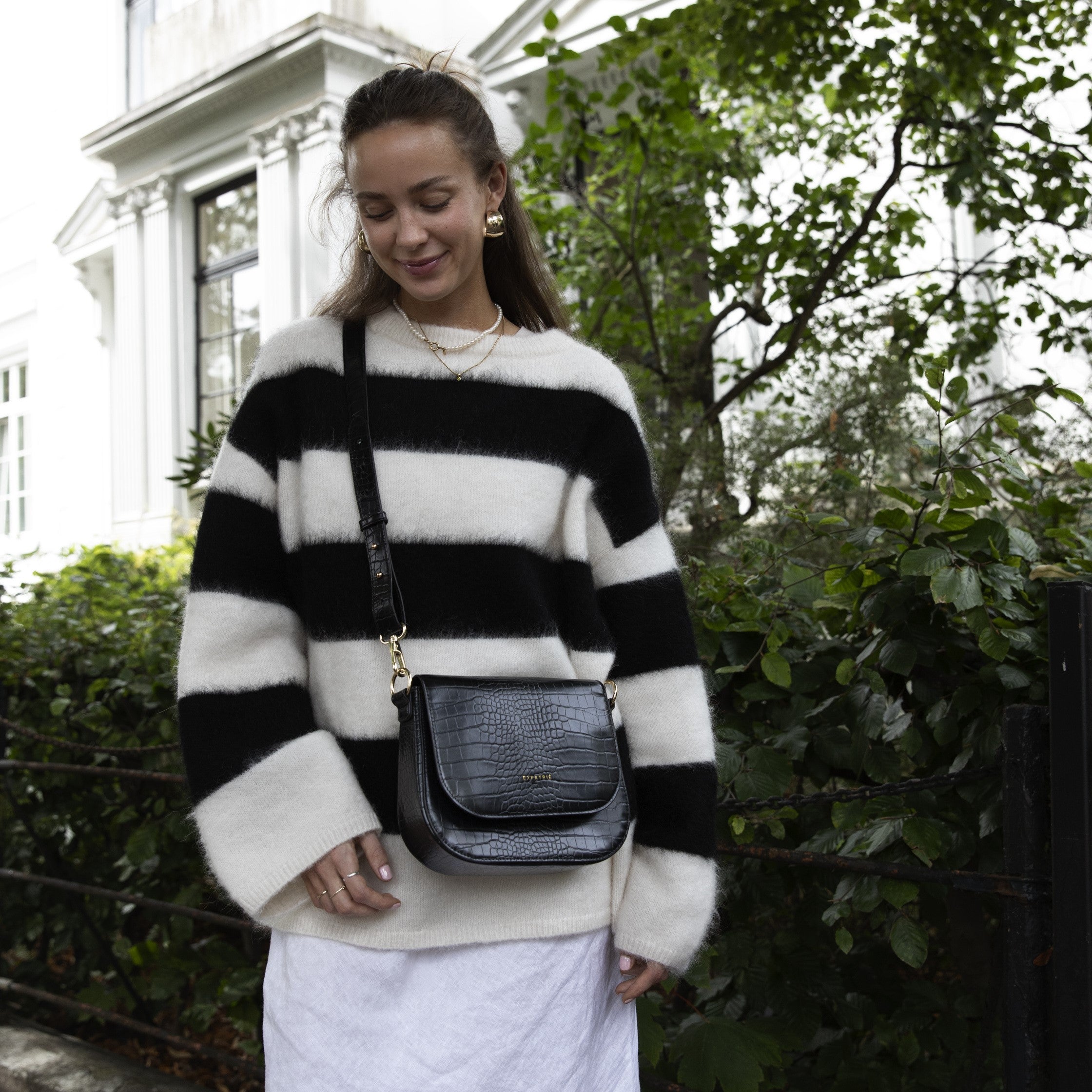 Frau trägt schwarze Saddle Bag Louise Medium mit Krokoprägung und goldenen Details über einen breit gestreiften schwarz-weißen Pullover