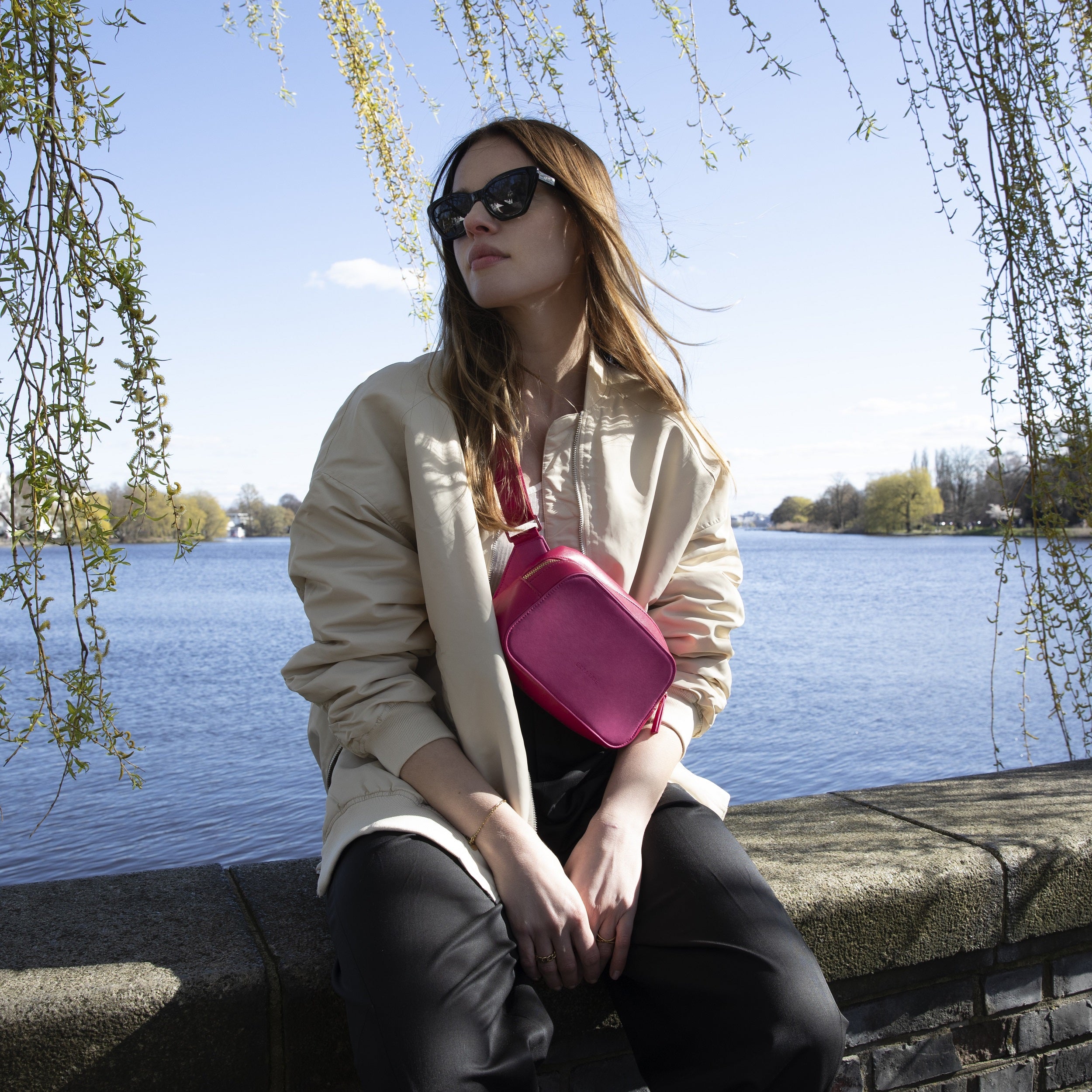 Frau sitzt am Wasser, trägt beige Jacke, schwarze Hose, Sonnenbrille und pinke Umhängetasche Fleur