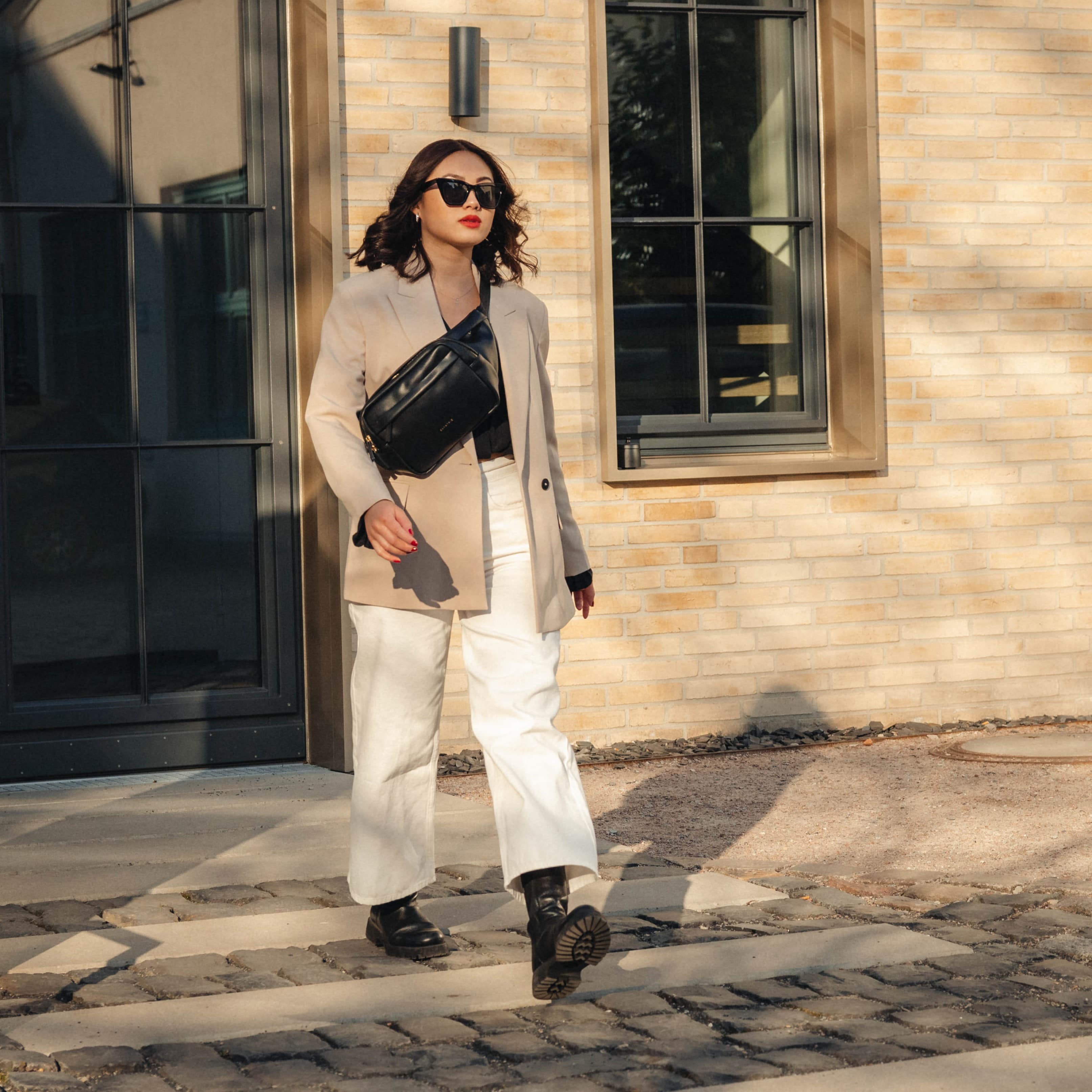 Frau trägt beige Blazer, weiße Hose, schwarze Stiefel und schwarze Bauchtasche Justine PU aus Leder