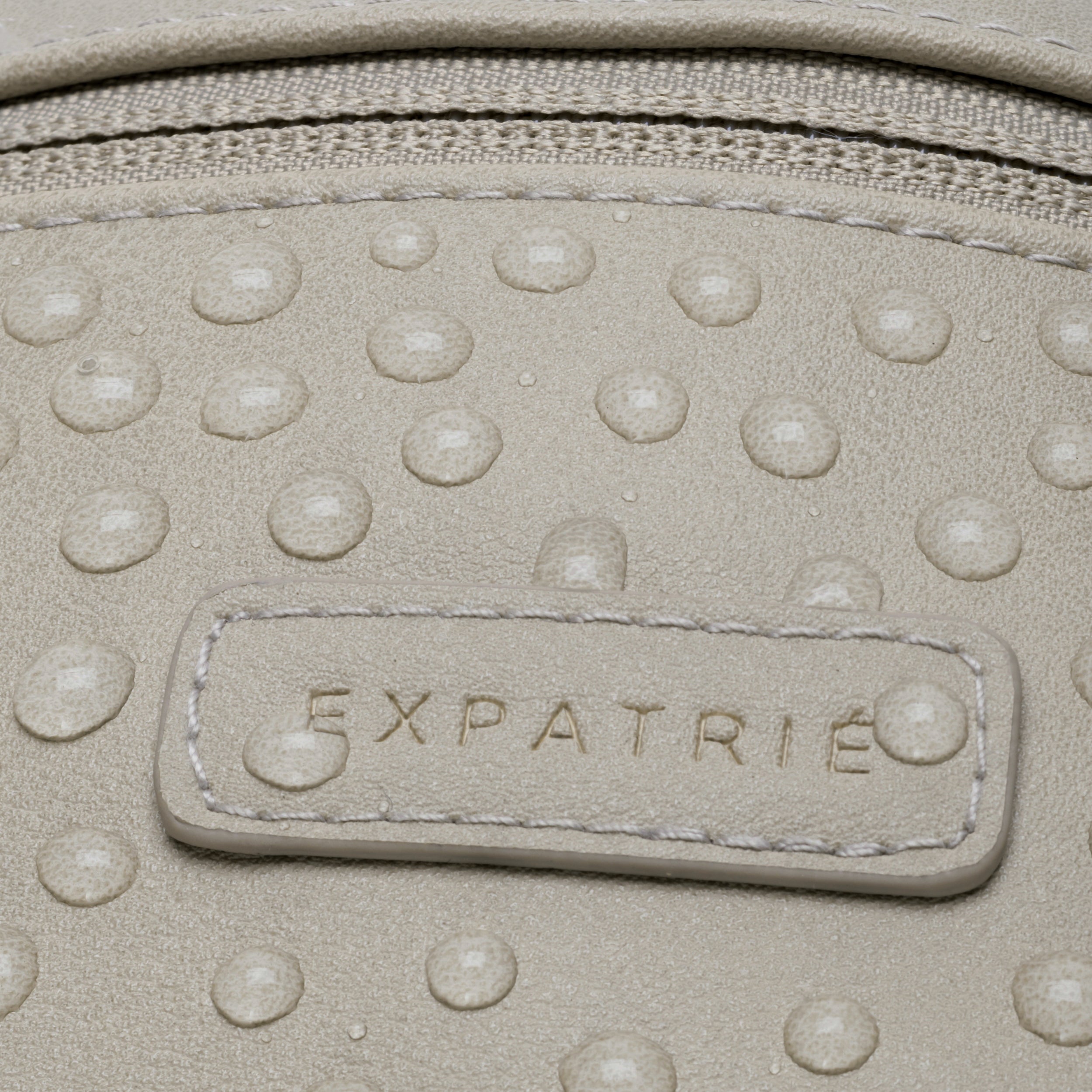 Wasserabweisende Oberfläche einer hellen Bauchtasche aus PU mit dem geprägten Markennamen 'Expatrié'