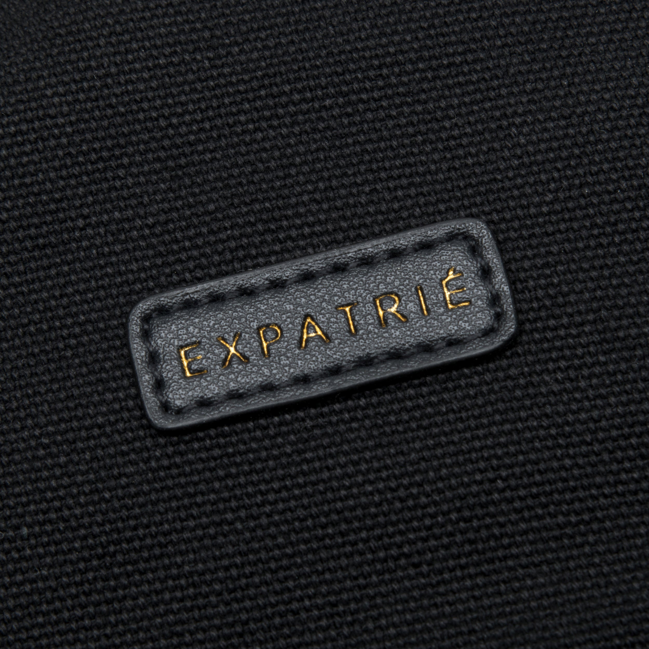 Schwarzes Canvas-Material mit gesticktem Lederetikett 'EXPATRIÉ' in Goldprägung