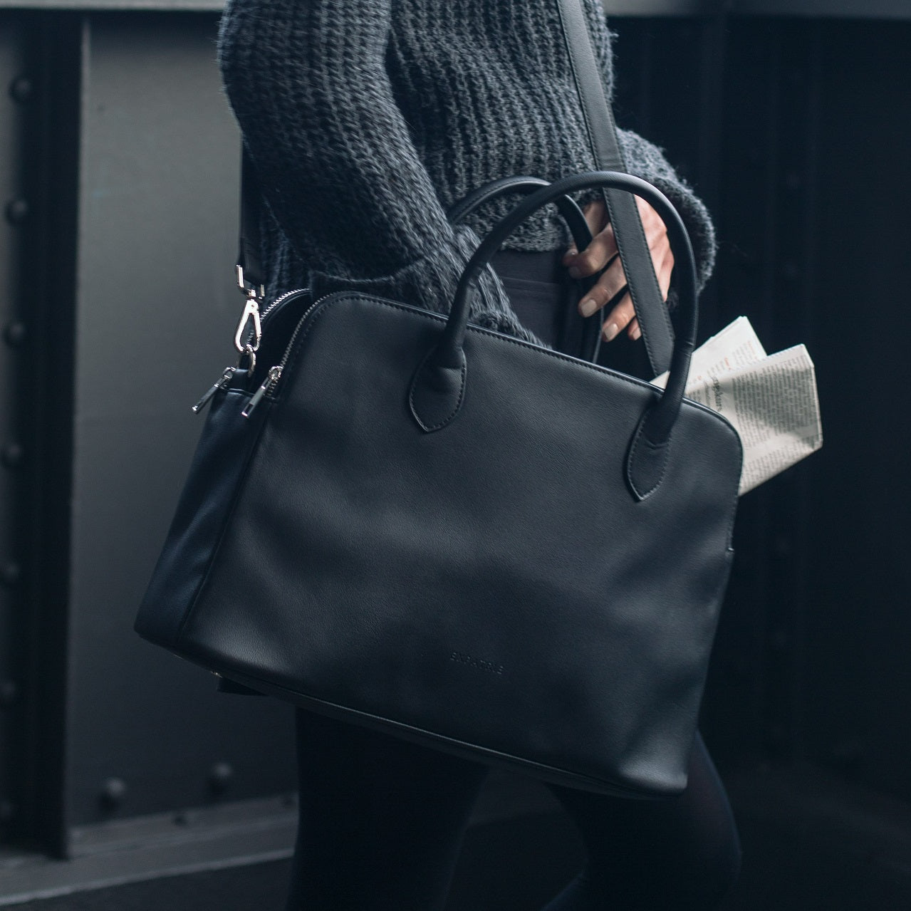 Schwarze Business-Handtasche Odette aus Leder mit Griffen und Handriemen, teilweise mit Zeitung gefüllt