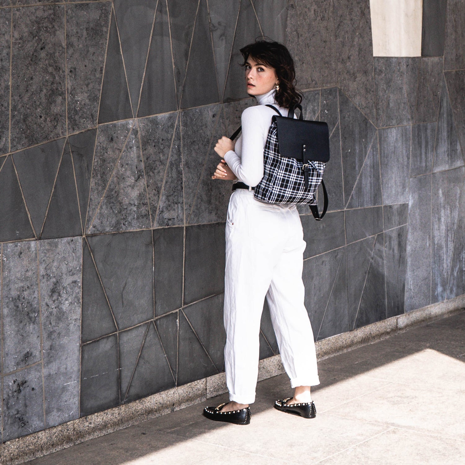 Frau mit schwarzem und weiß kariertem Canvas-Leder-Rucksack Anouk, trägt weißen Rollkragenpullover und weiße Hose