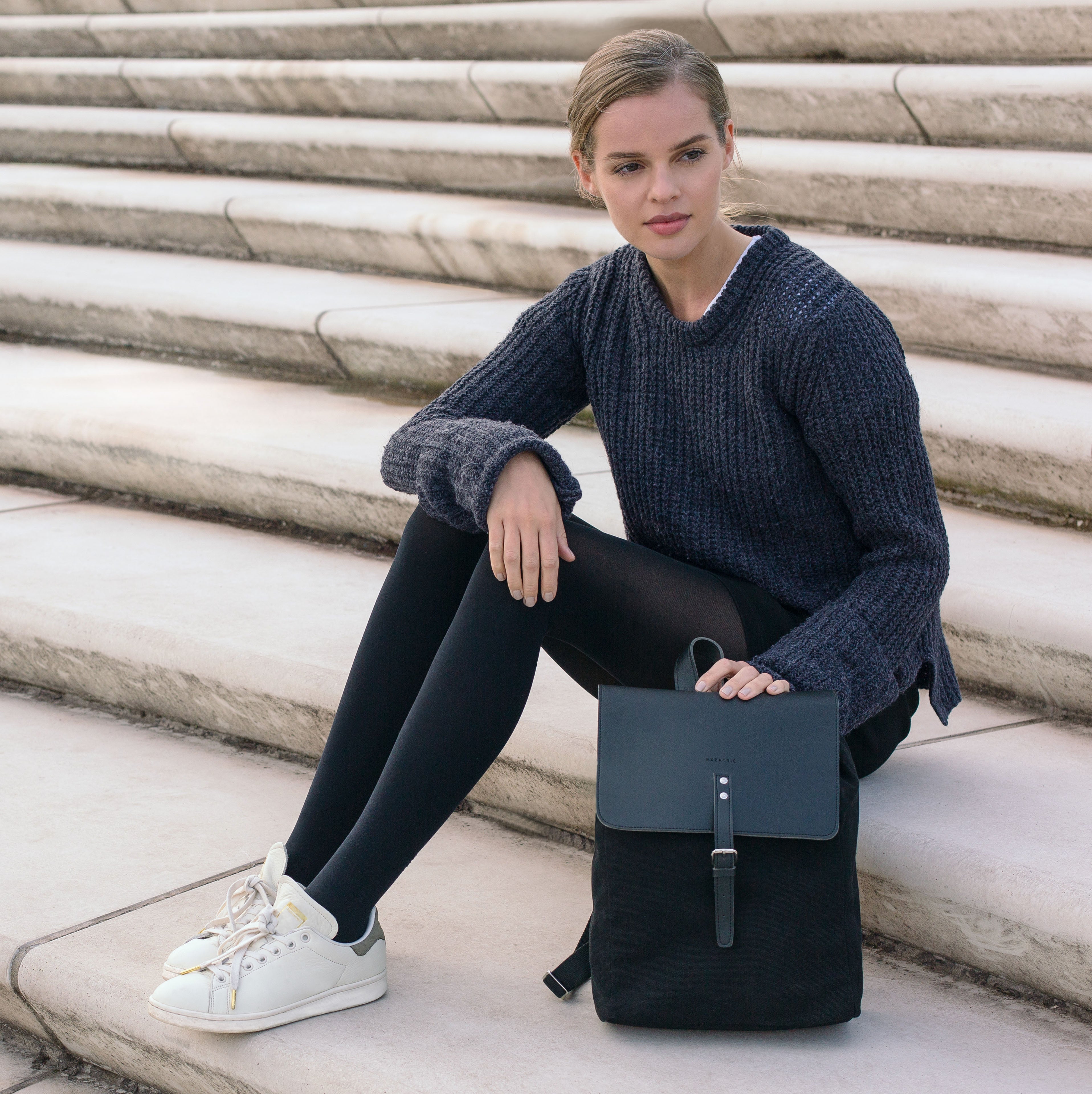 Frau sitzt auf Marmortreppe mit schwarzem Canvas-Leder-Rucksack Anouk und trägt dunklen Pullover, Strumpfhose und weiße Sneaker