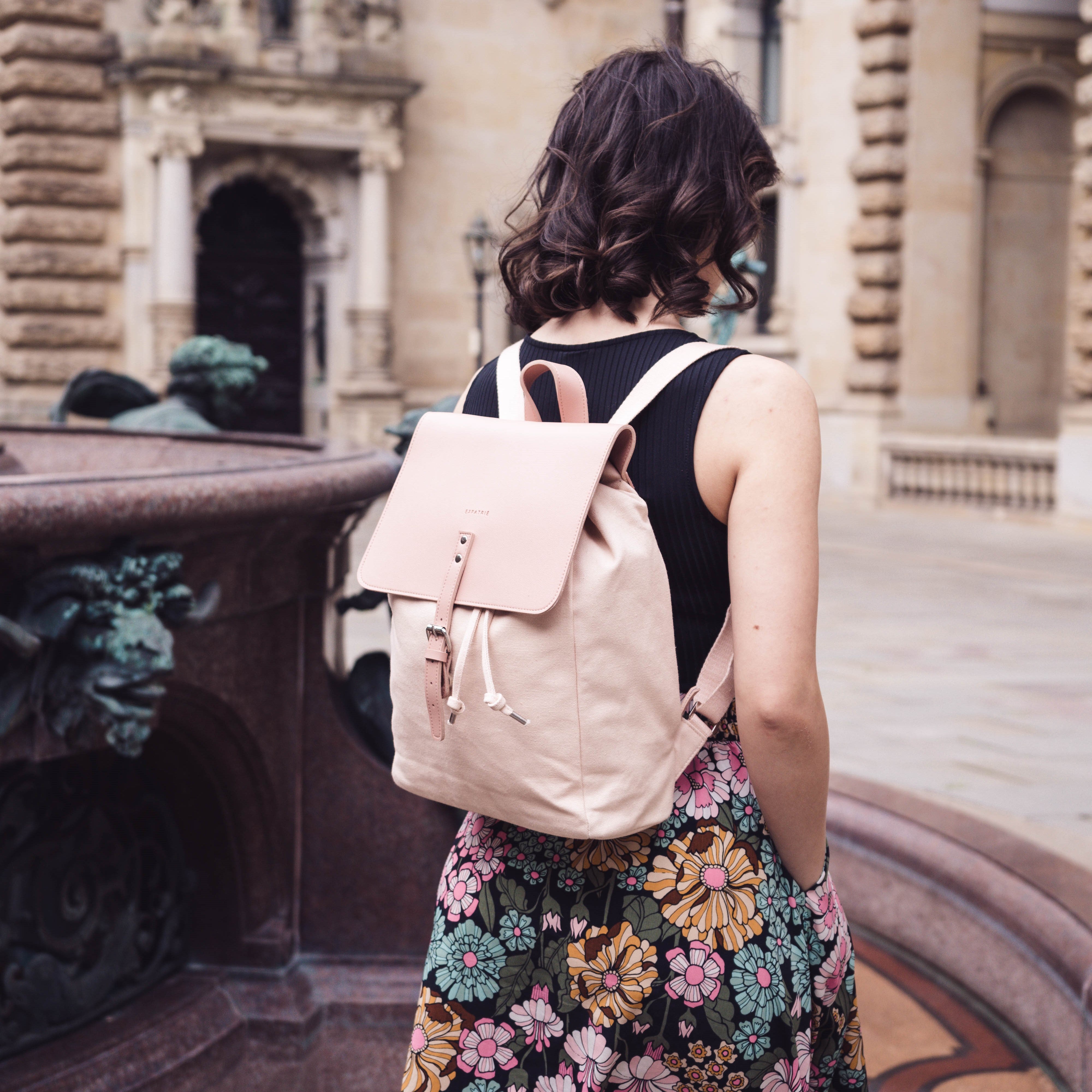 Frau mit schwarzem Top und Blumenrock trägt rosa Canvas-Rucksack Anouk an historischem Gebäuderand