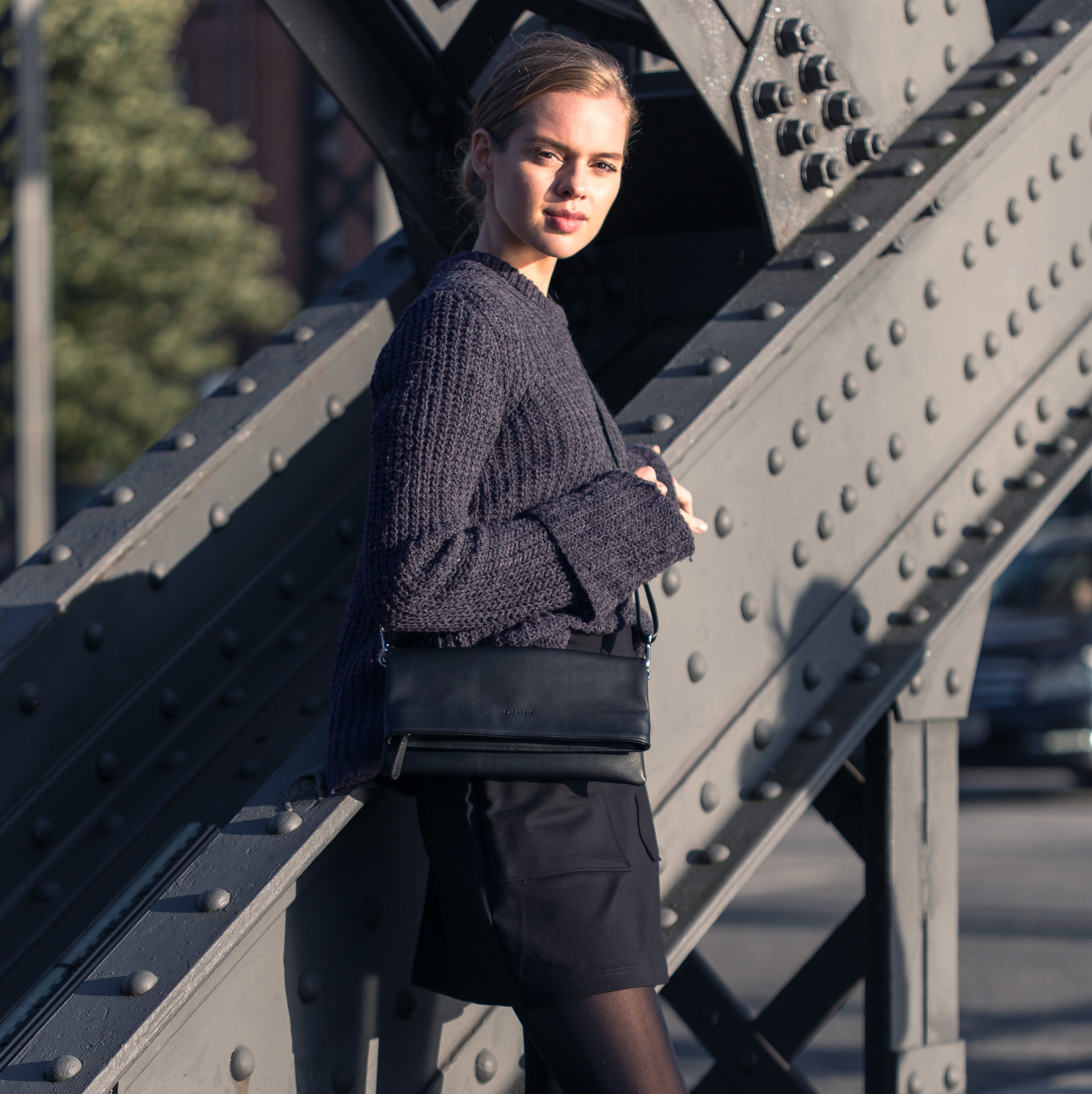 Frau in dunklem Pullover mit schwarzer Clutch Marie Tasche an grauer Metallstruktur