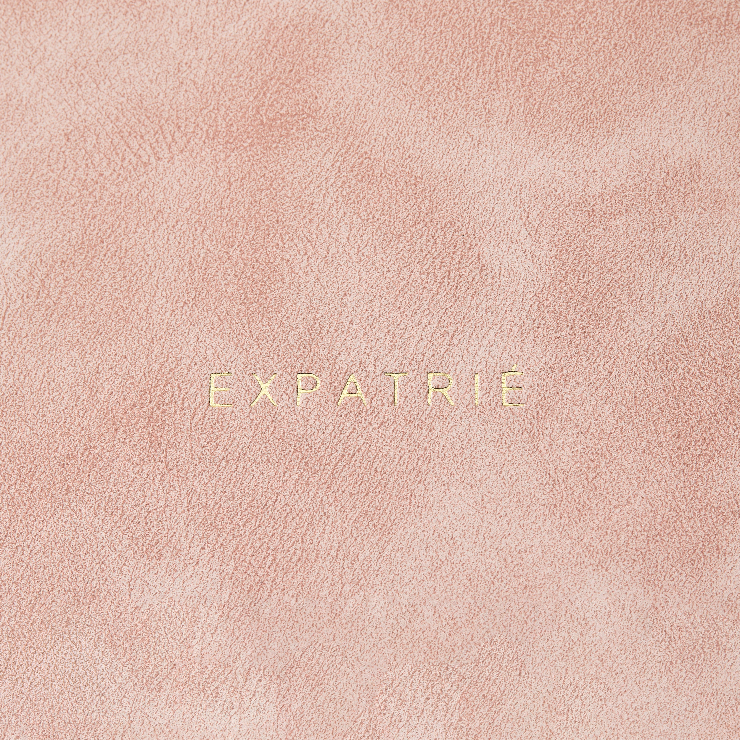 Nahe Aufnahme der goldenen Prägung 'EXPATRIÉ' auf einer rosa-nude Lederoberfläche der Clutch Isabelle