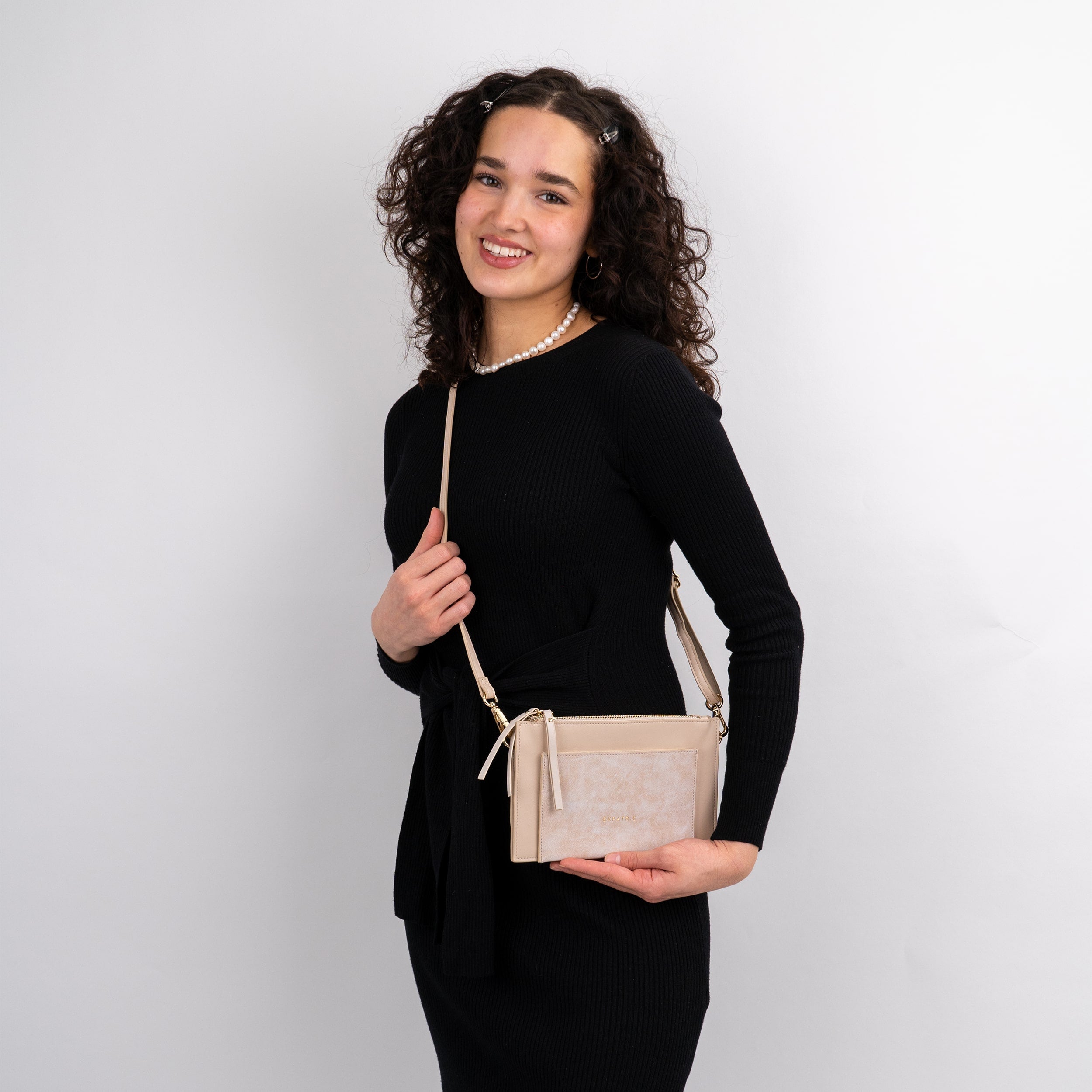 Frau mit lockigem Haar trägt beigefarbene Clutch 'Isabelle' mit Schulterriemen, schwarzes Kleid und Perlenkette