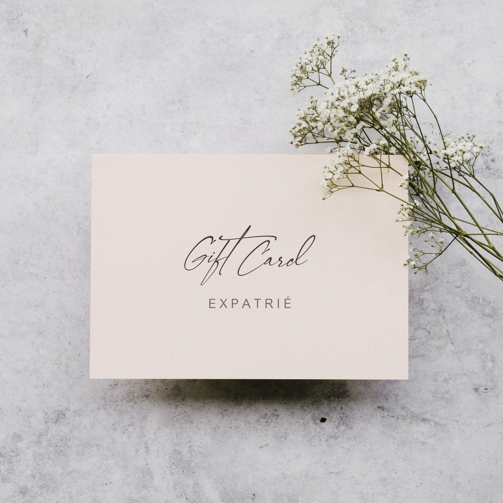 Geschenkgutschein mit der Aufschrift 'Gift Card Expatrié' neben weißen Blüten auf grauem Untergrund