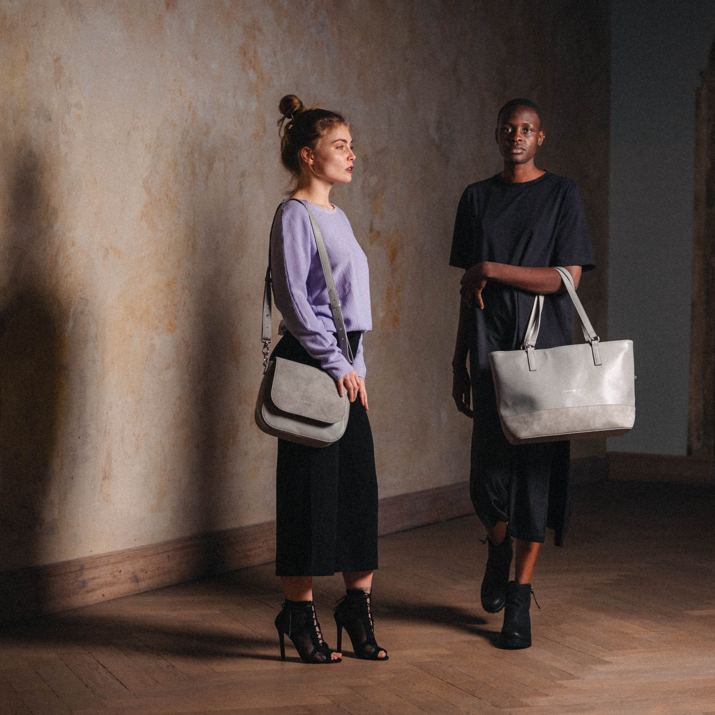 Zwei Frauen stehen in einem Innenraum mit holzboden, eine trägt eine graue Saddle Bag Louise Large über der Schulter
