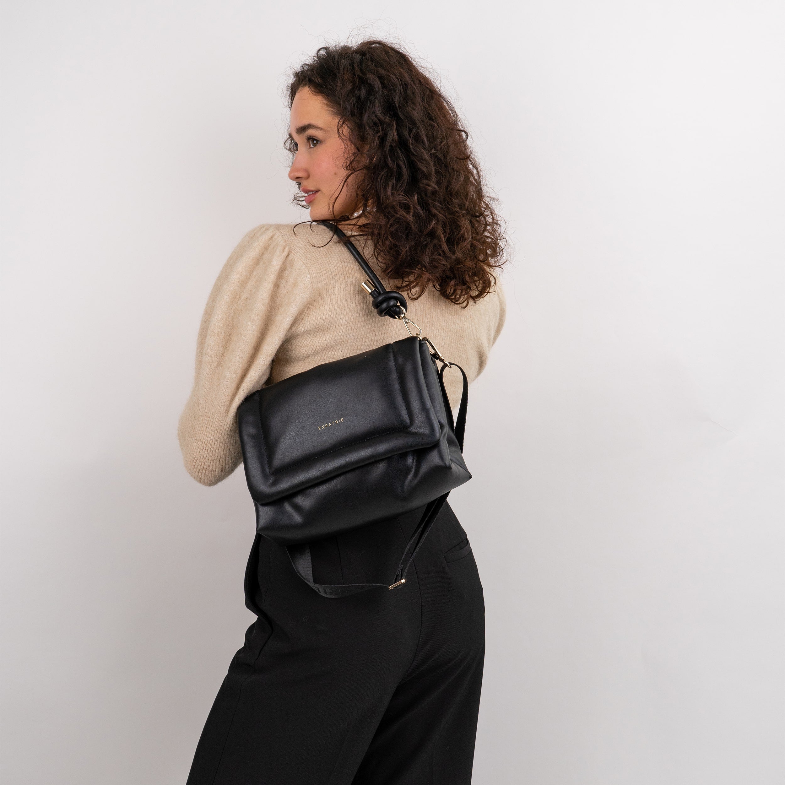 Frau trägt Umhängetasche Zoe Medium in Schwarz über Schulter, beige Pullover und schwarze Hose