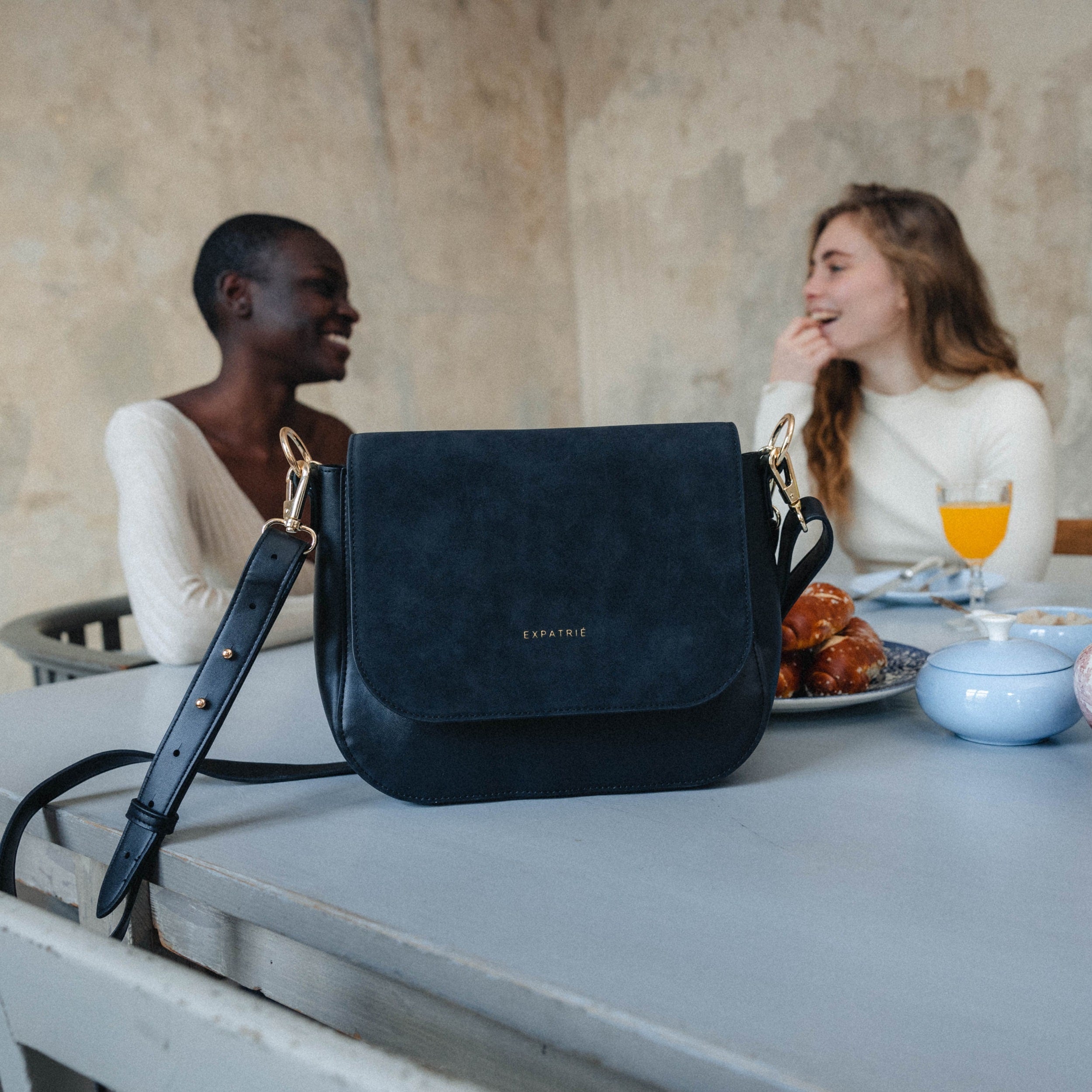 Große schwarze Saddle Bag Louise von Expatrié auf einem Tisch mit Frühstück und zwei Frauen im Hintergrund