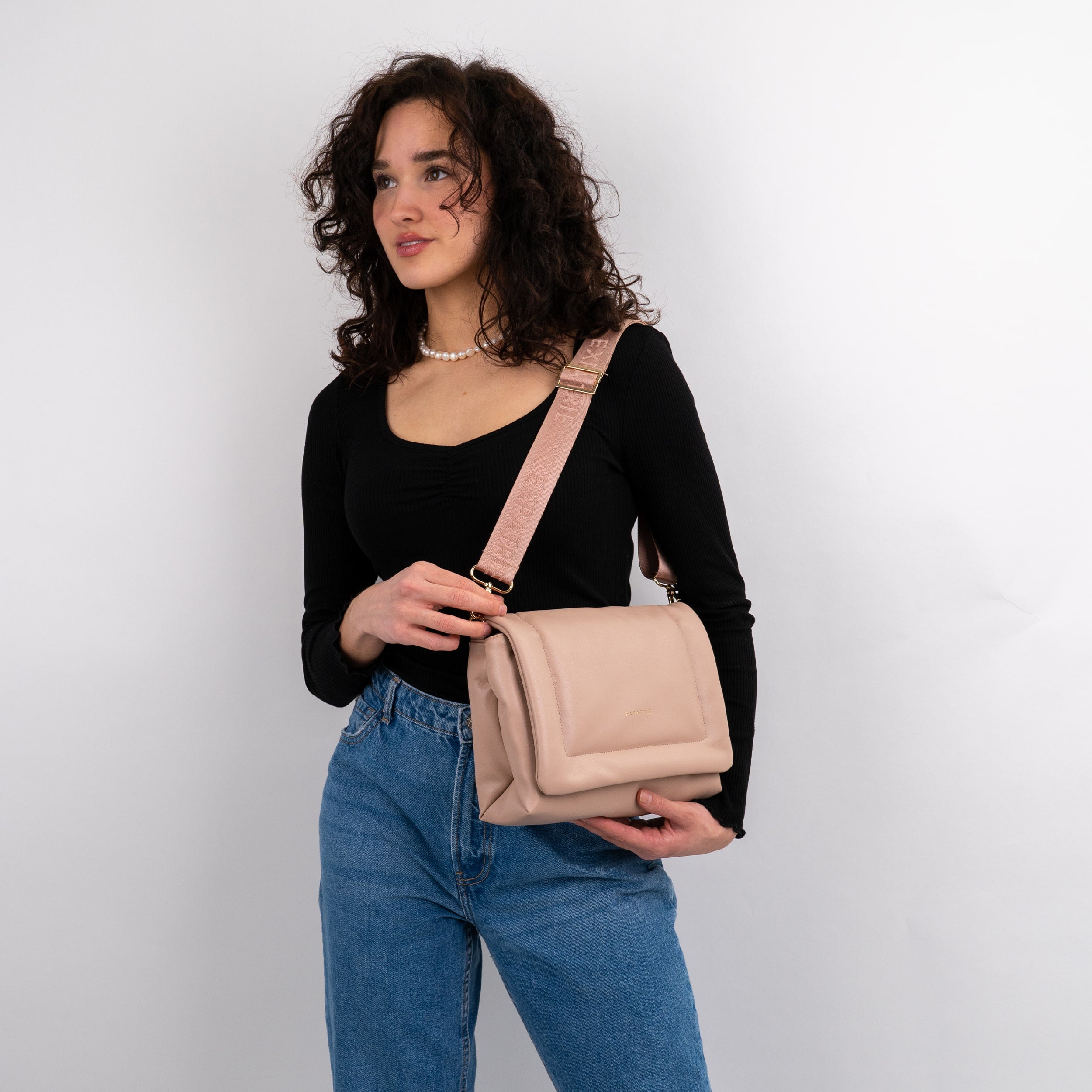 Frau trägt beige Umhängetasche Zoe Medium mit breitem Riemen, schwarze Bluse und blaue Jeans