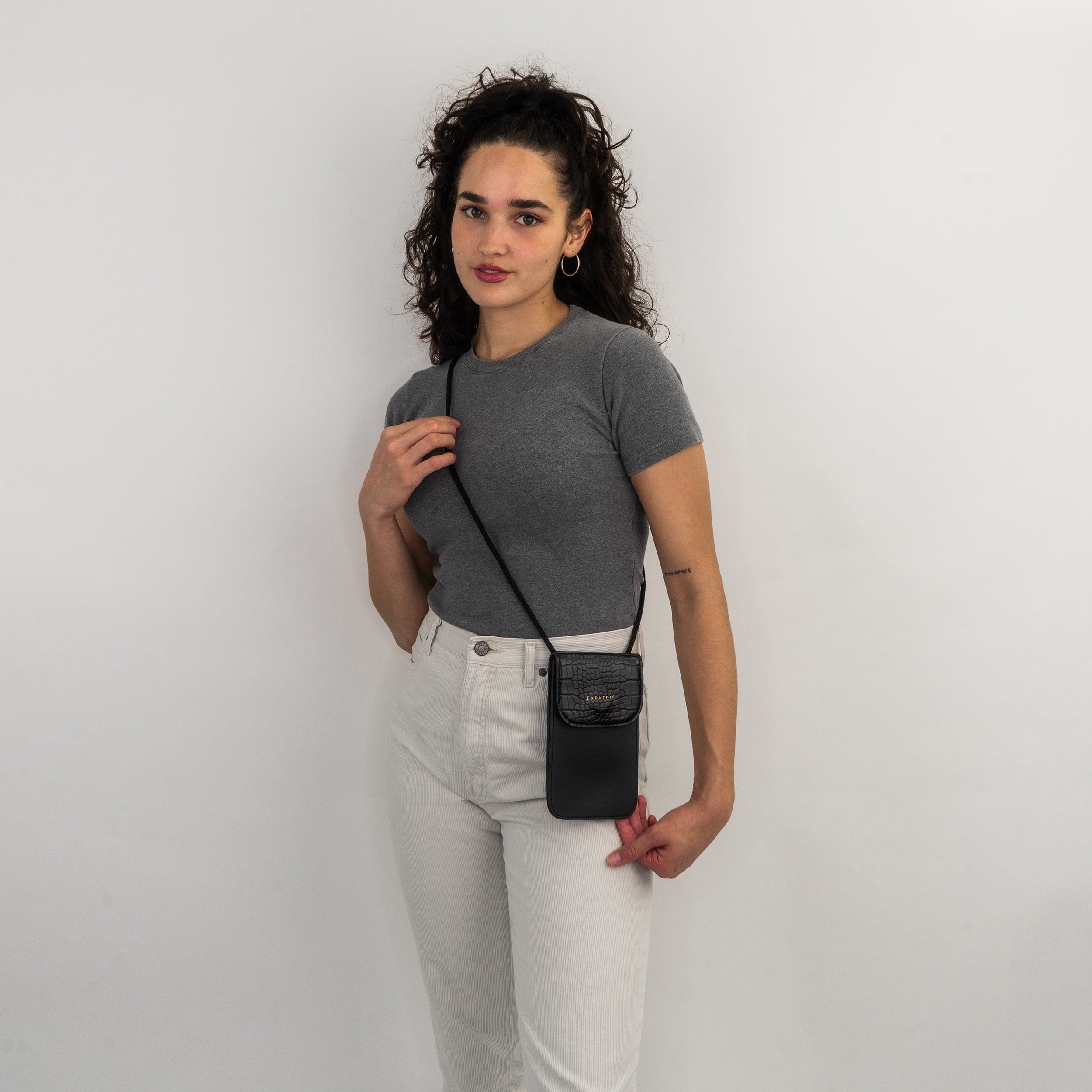 Frau trägt schwarze Handy Umhängetasche Amelie mit Kroko-Muster und goldfarbenem Logo, dazu graues Shirt und weiße Hose