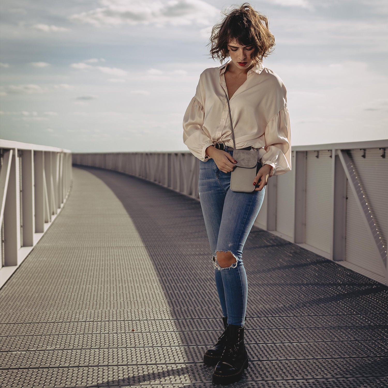 Frau mit lockigem Haar trägt beige Bluse und enge, zerrissene blaue Jeans mit grauer Umhängetasche Amelie auf einer Brücke