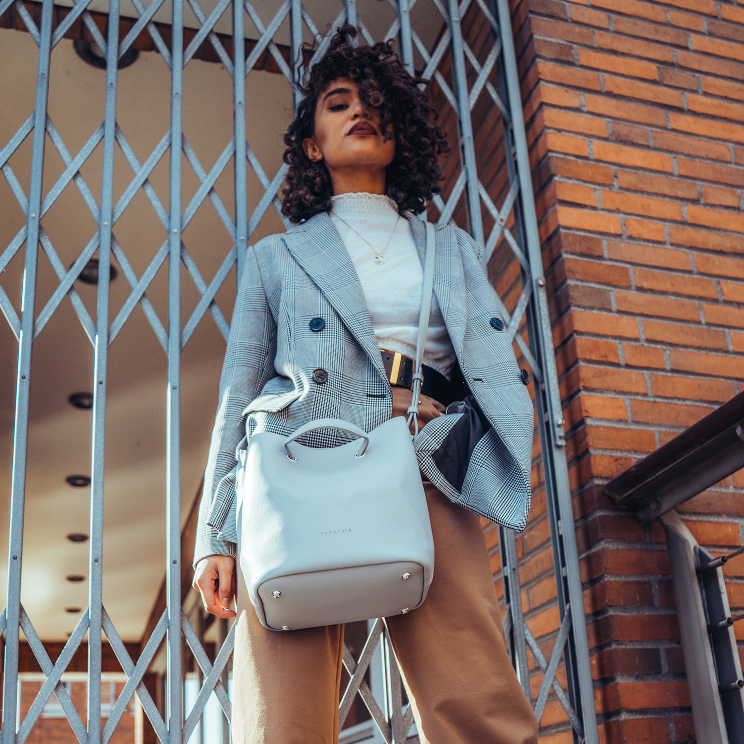 Frau mit lockigem Haar trägt graue Beuteltasche Élodie, hellgrauer Blazer und beige Hose