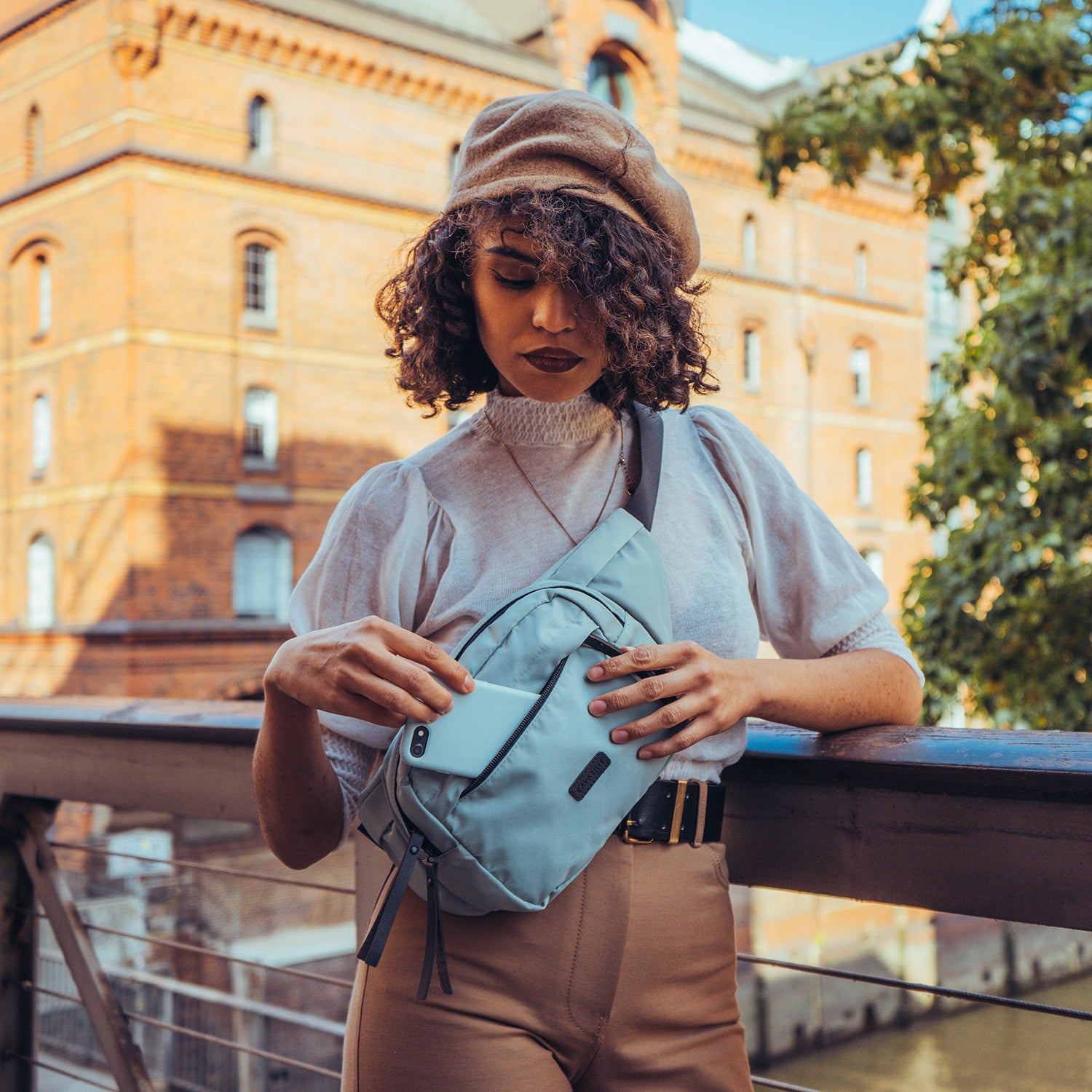 Frau mit lockigem Haar und Baskenmütze entnimmt Handy aus mintgrüner Bauchtasche Justine aus Nylon