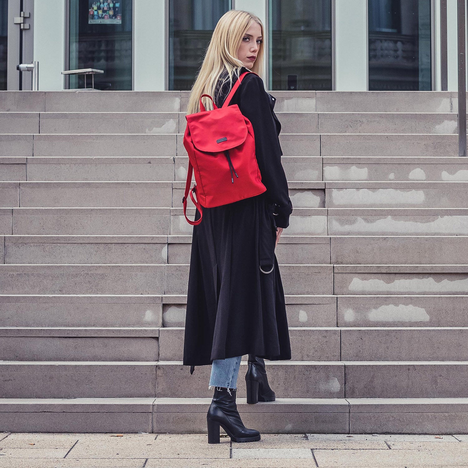 Frau mit langem schwarzem Mantel und rotem Nylon Rucksack Clara steht vor Treppe