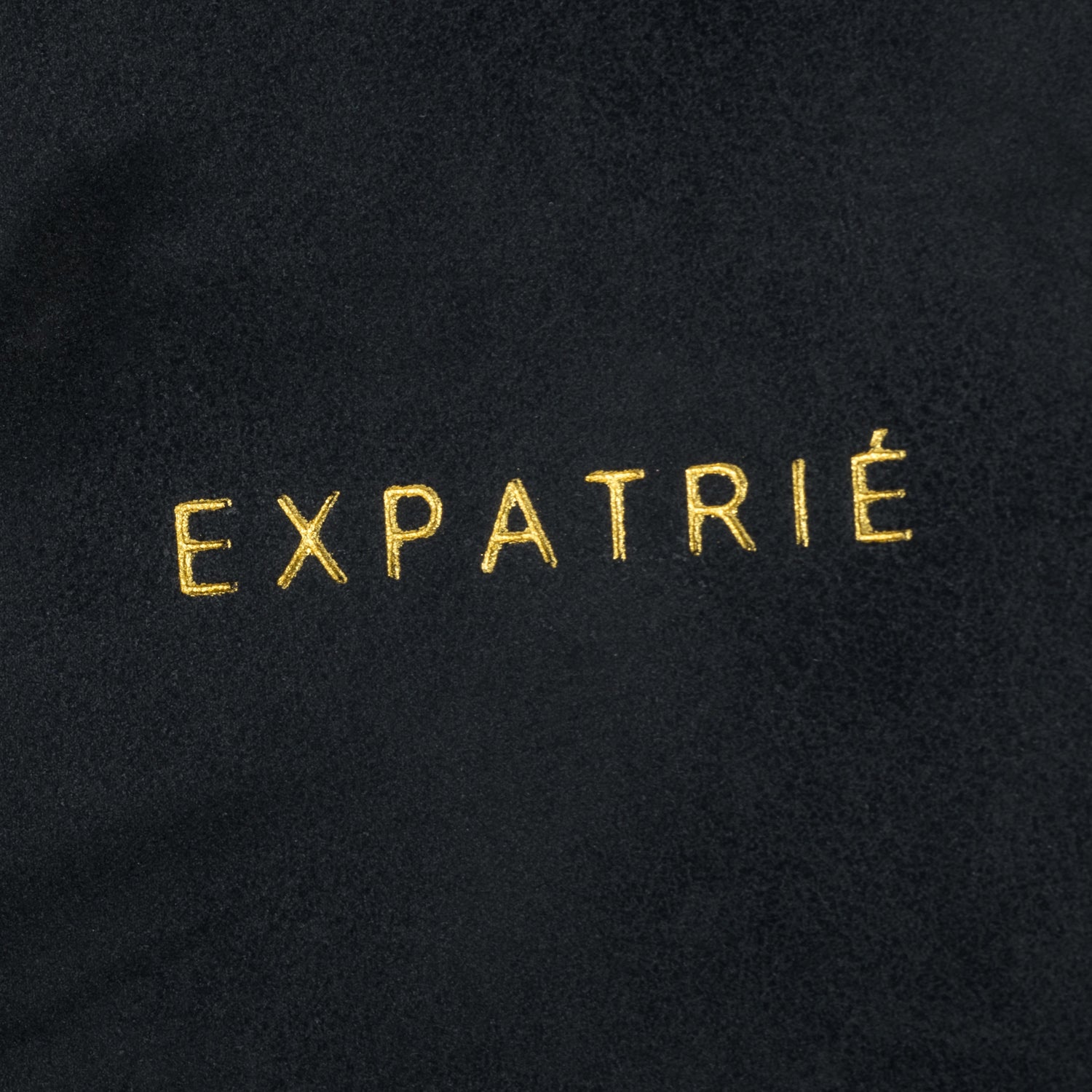 Schwarzes Kunstleder mit goldener Stickerei des Schriftzugs 'EXPATRIÉ'