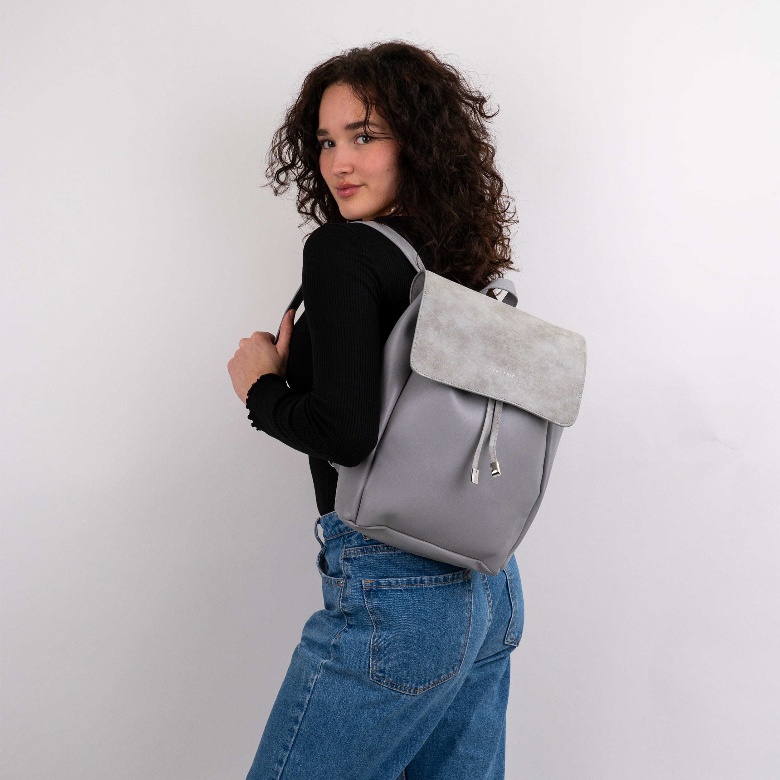 Frau trägt grauen Kunstleder Rucksack Estelle mit Umschlag und Kordelzug, schwarze Longsleeve und blaue Jeans