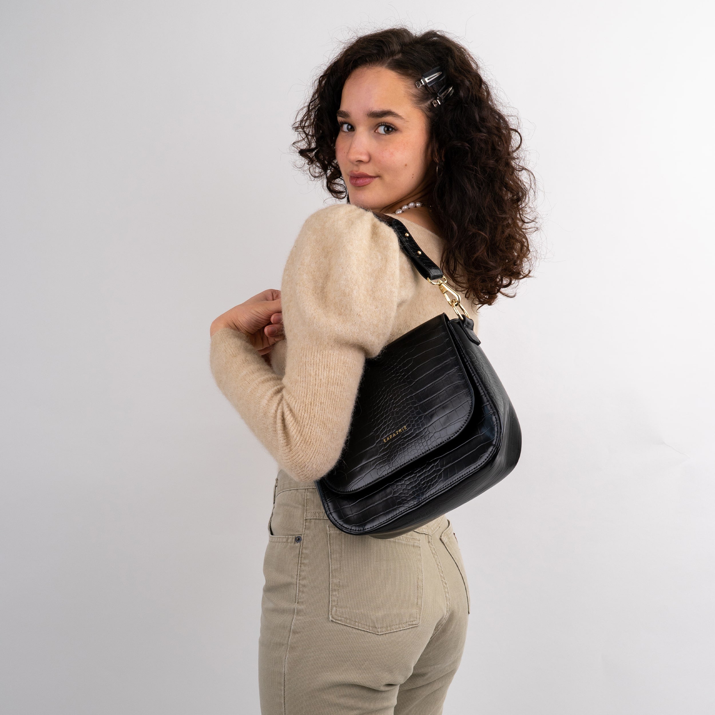 Frau mit lockigem Haar trägt schwarze Saddle Bag Louise Large Schultertasche aus Kroko-Leder