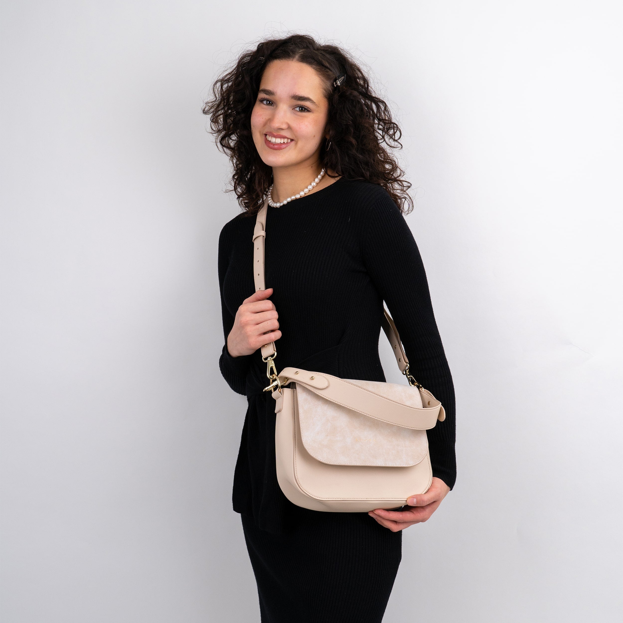 Frau mit lockigem Haar trägt große beige Saddle Bag Louise aus Leder über der Schulter, schwarzes Kleid